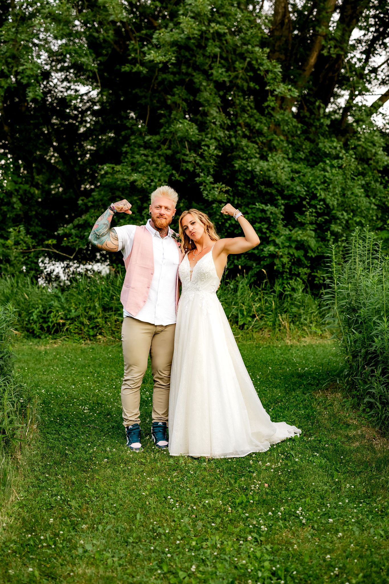 Bride and groom flexing muscles together outdoors on green grass, wearing wedding attire with trees in background.