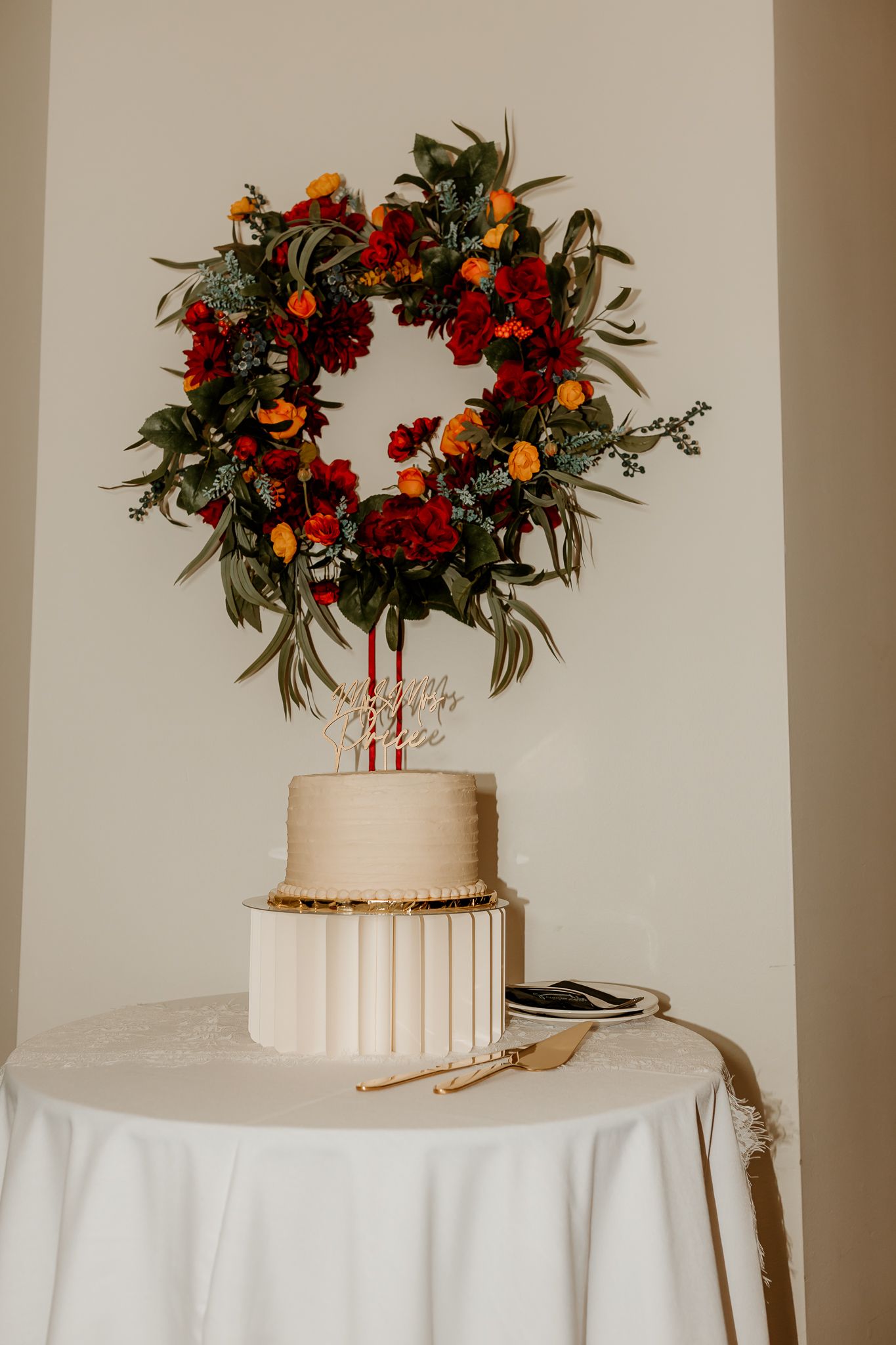 Two-tier white wedding cake with gold accents topped by a circular wreath of red and orange flowers on a draped table with cake server.