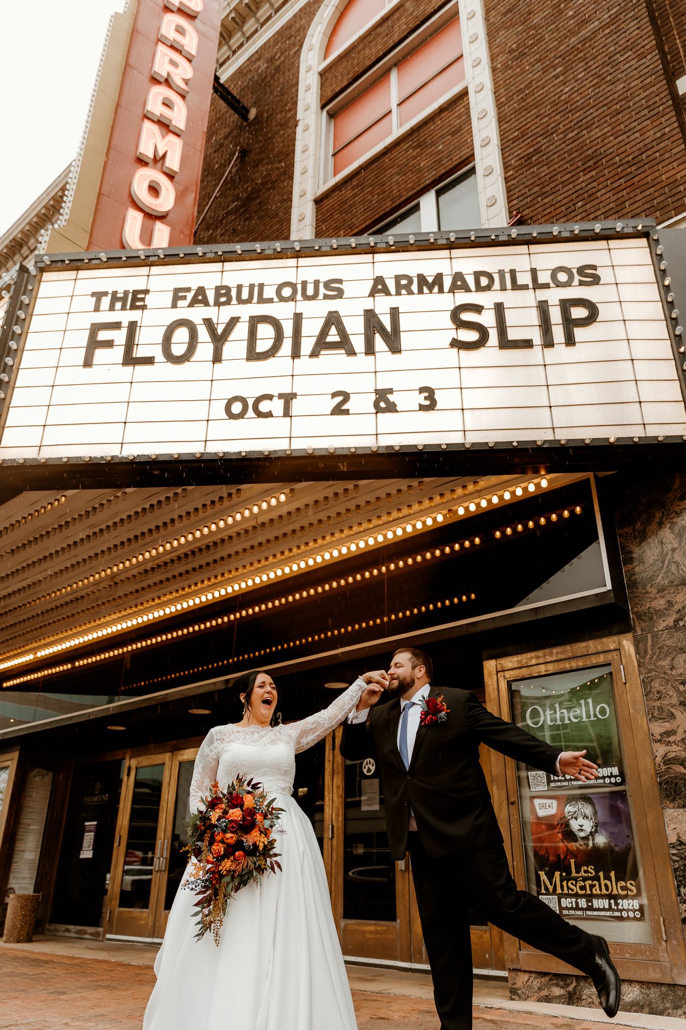 Bride in a white gown with an orange bouquet and groom in a black suit dance joyfully beneath a vintage theater marquee.