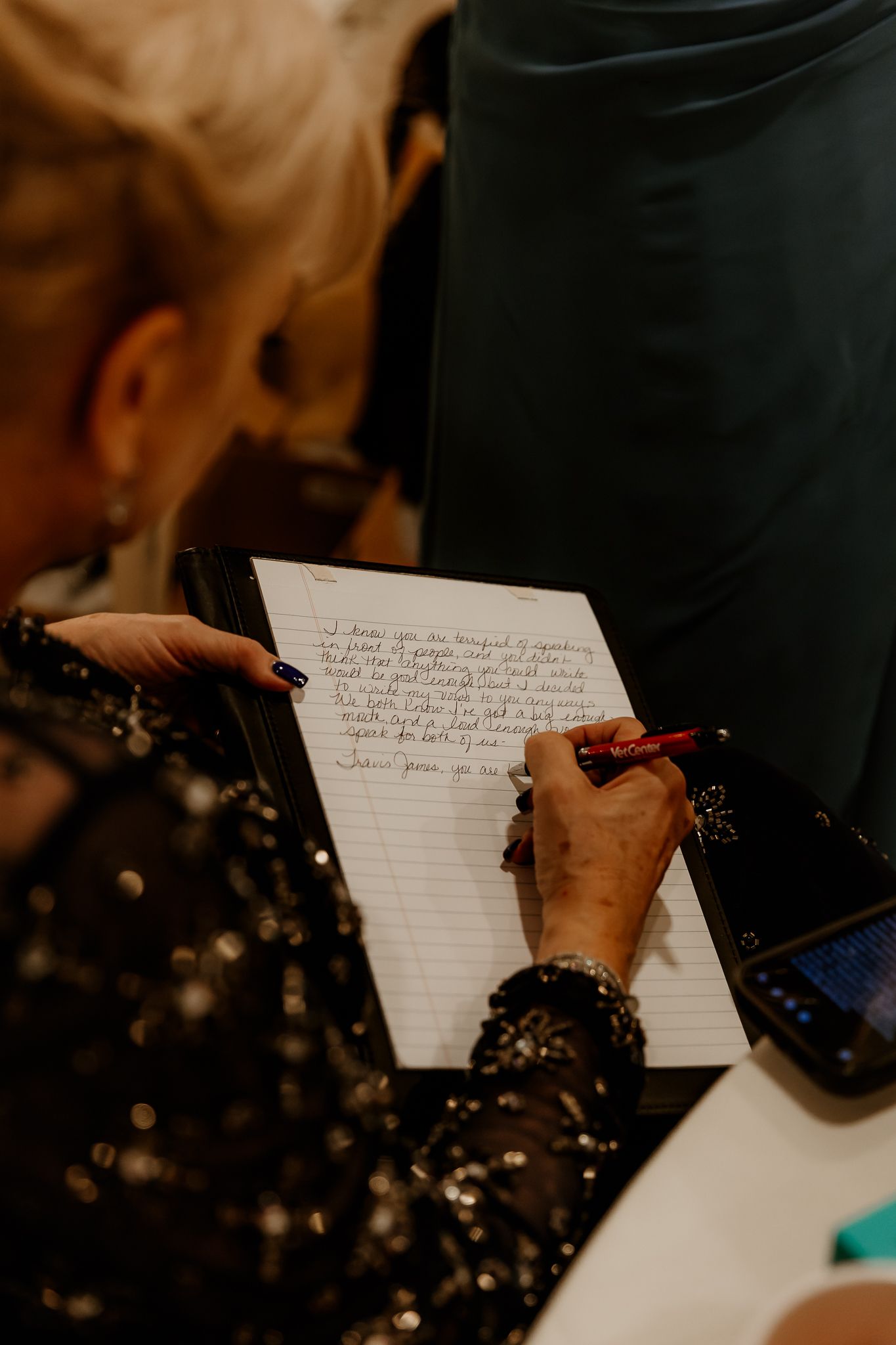 A woman in a sparkly dark dress with painted nails writes a handwritten message in a guest book with a red pen.