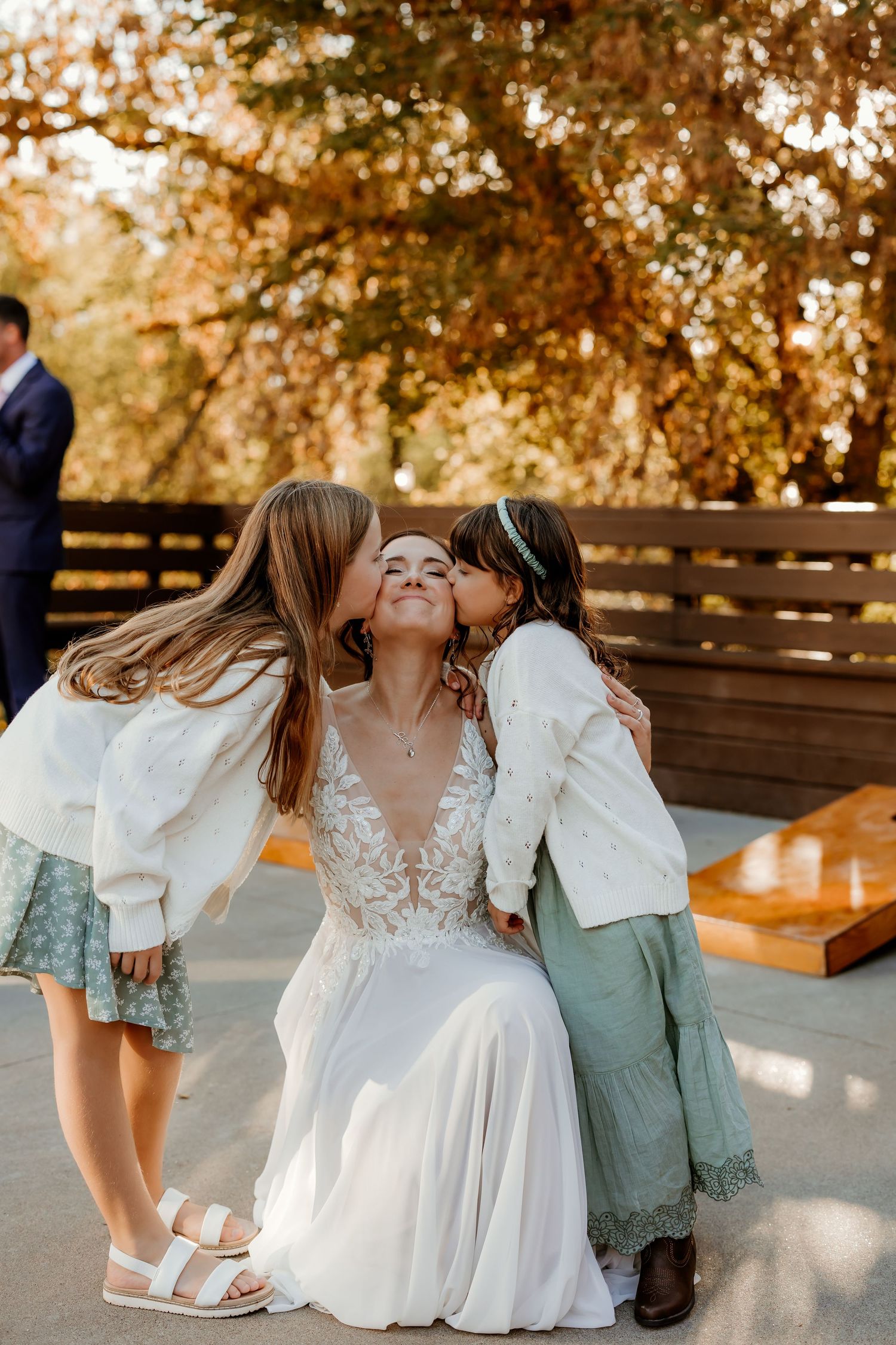 Bride in a lace wedding dress smiles while two young girls in cardigans and skirts kiss her cheeks outdoors beneath autumn trees.