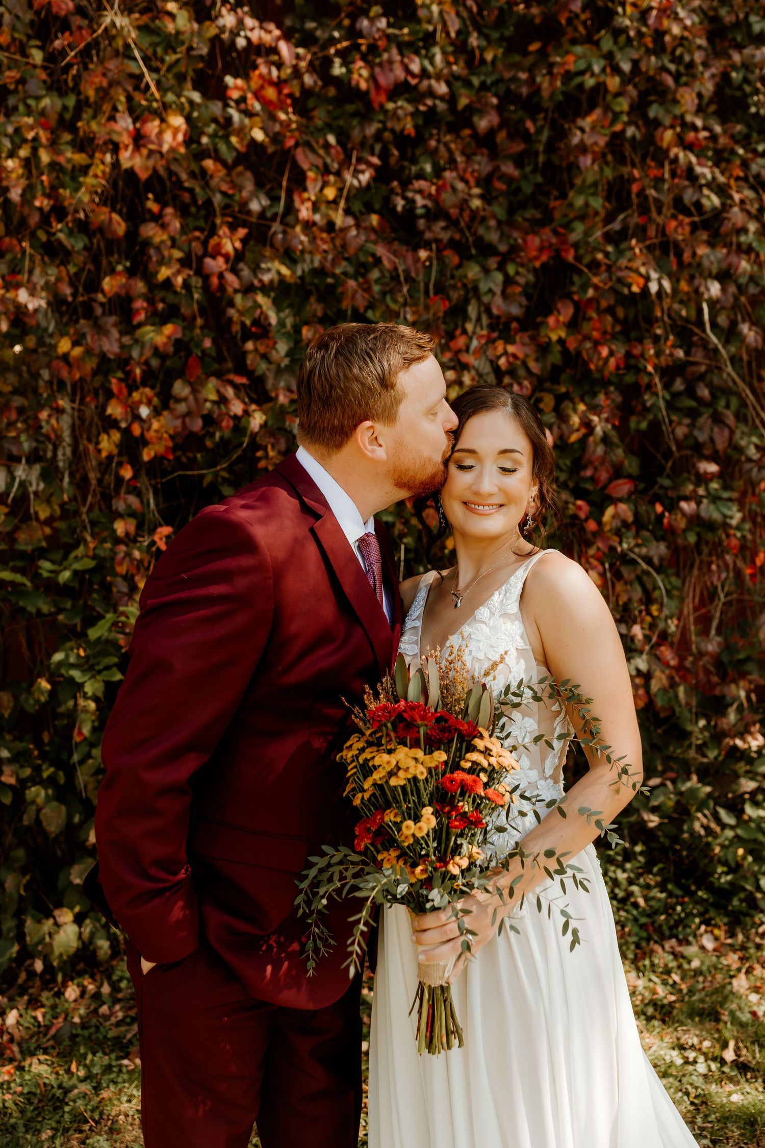 Groom in a burgundy suit kisses smiling bride in a white gown on the forehead as she holds a fall bouquet before ivy-covered autumn foliage.