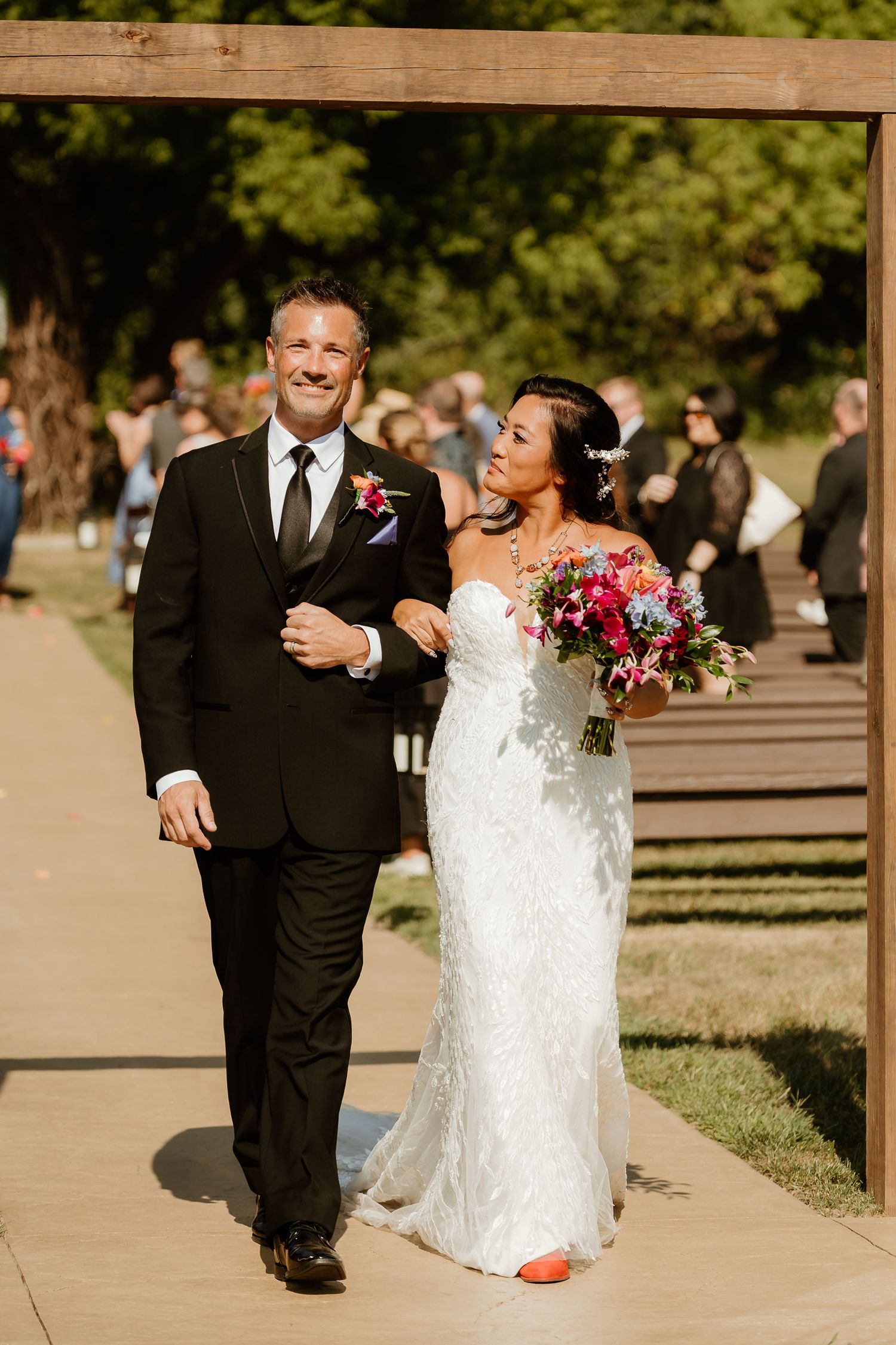 Smiling bride in white lace gown with colorful bouquet walks arm-in-arm with groom in black suit at outdoor wedding.