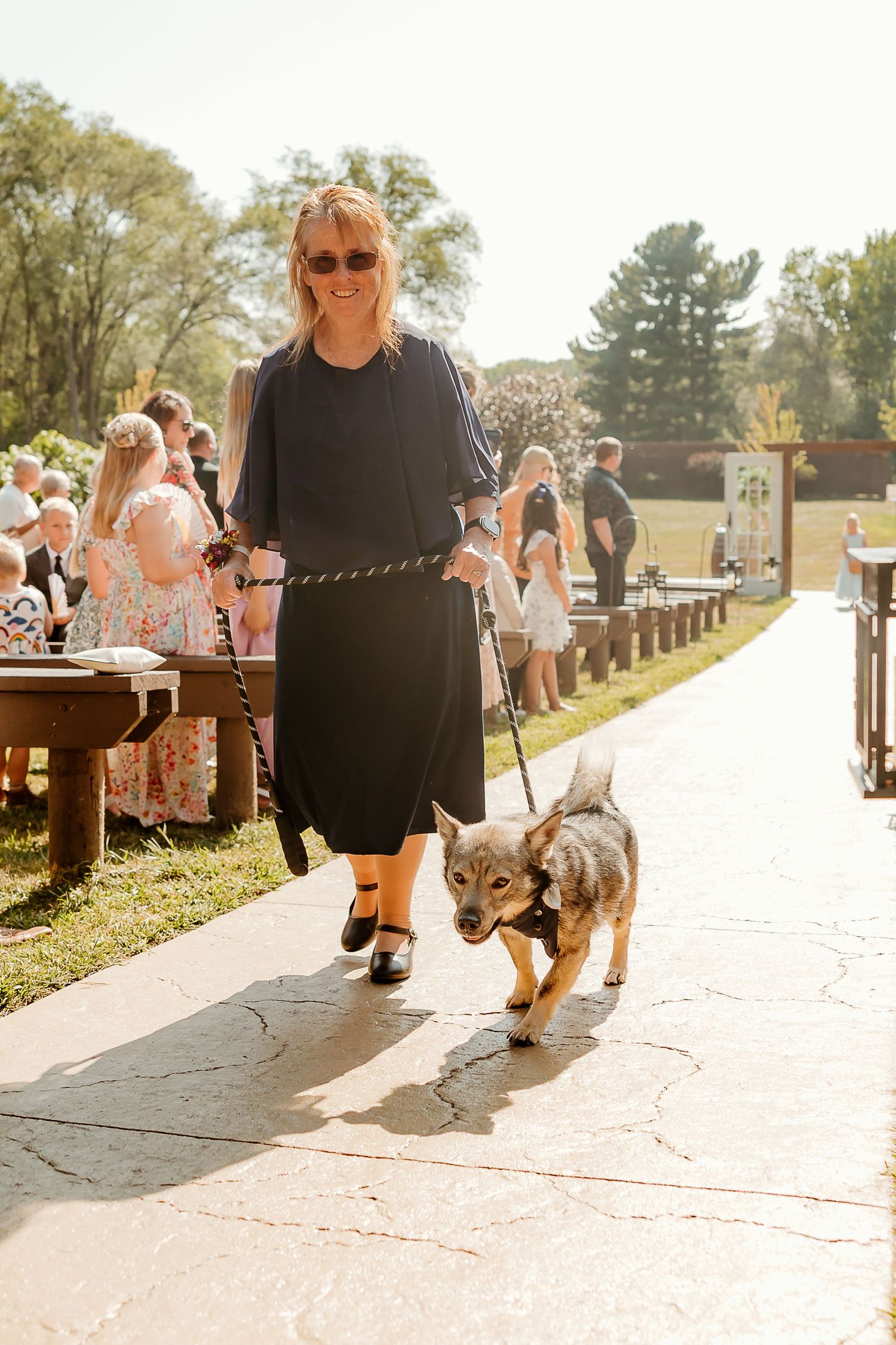 Woman in black dress walks small dog on leash down outdoor wedding aisle on sunny day.