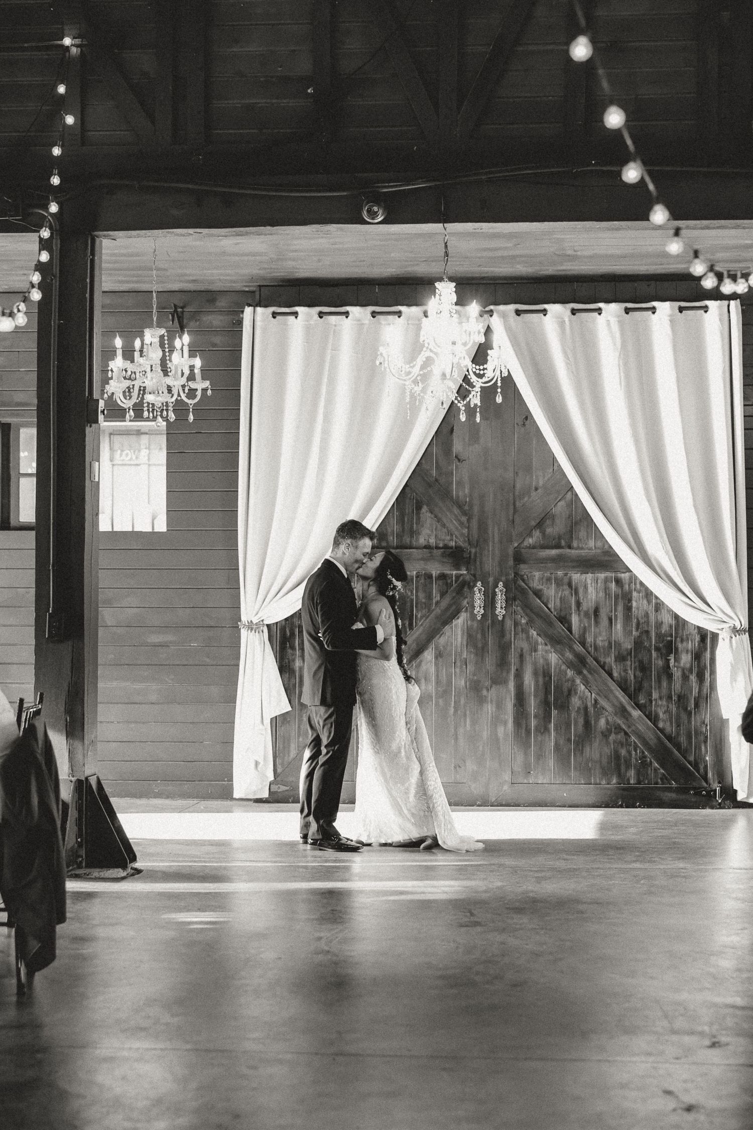 Black and white photo of a bride and groom sharing their first dance in a rustic barn venue with chandeliers.