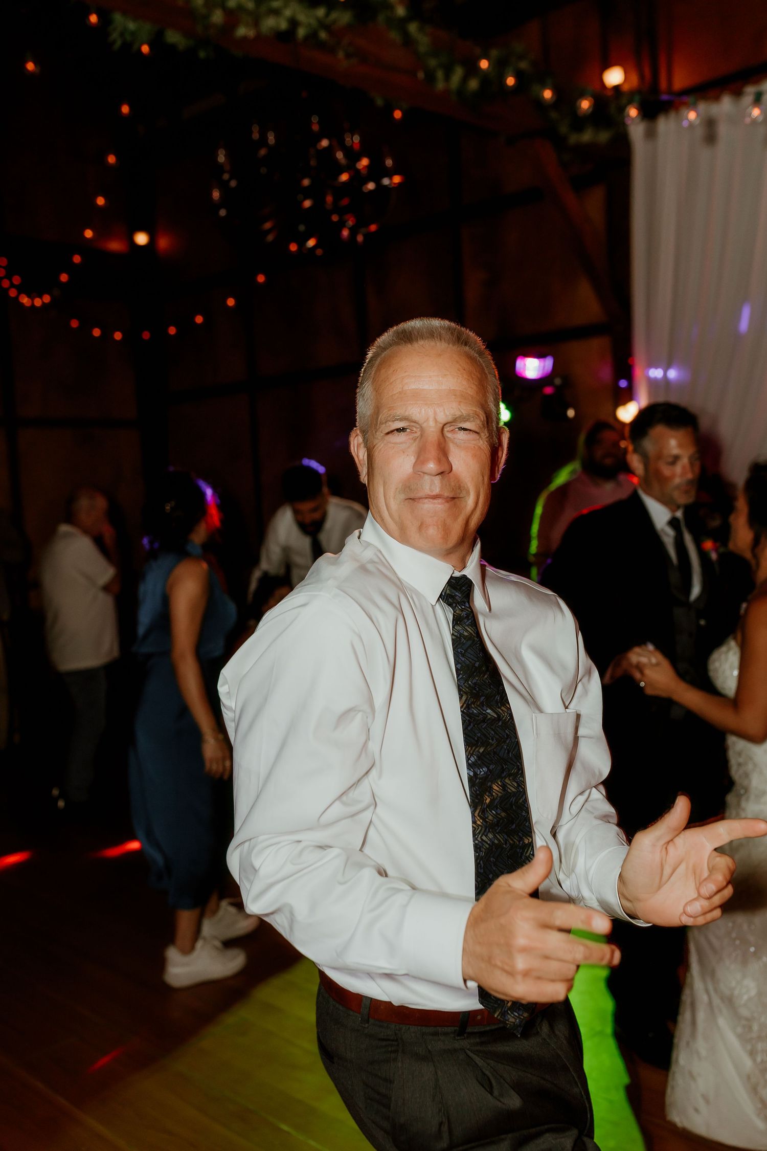 Middle-aged man in white dress shirt and dark tie smiling on a wedding reception dance floor with colorful lights.
