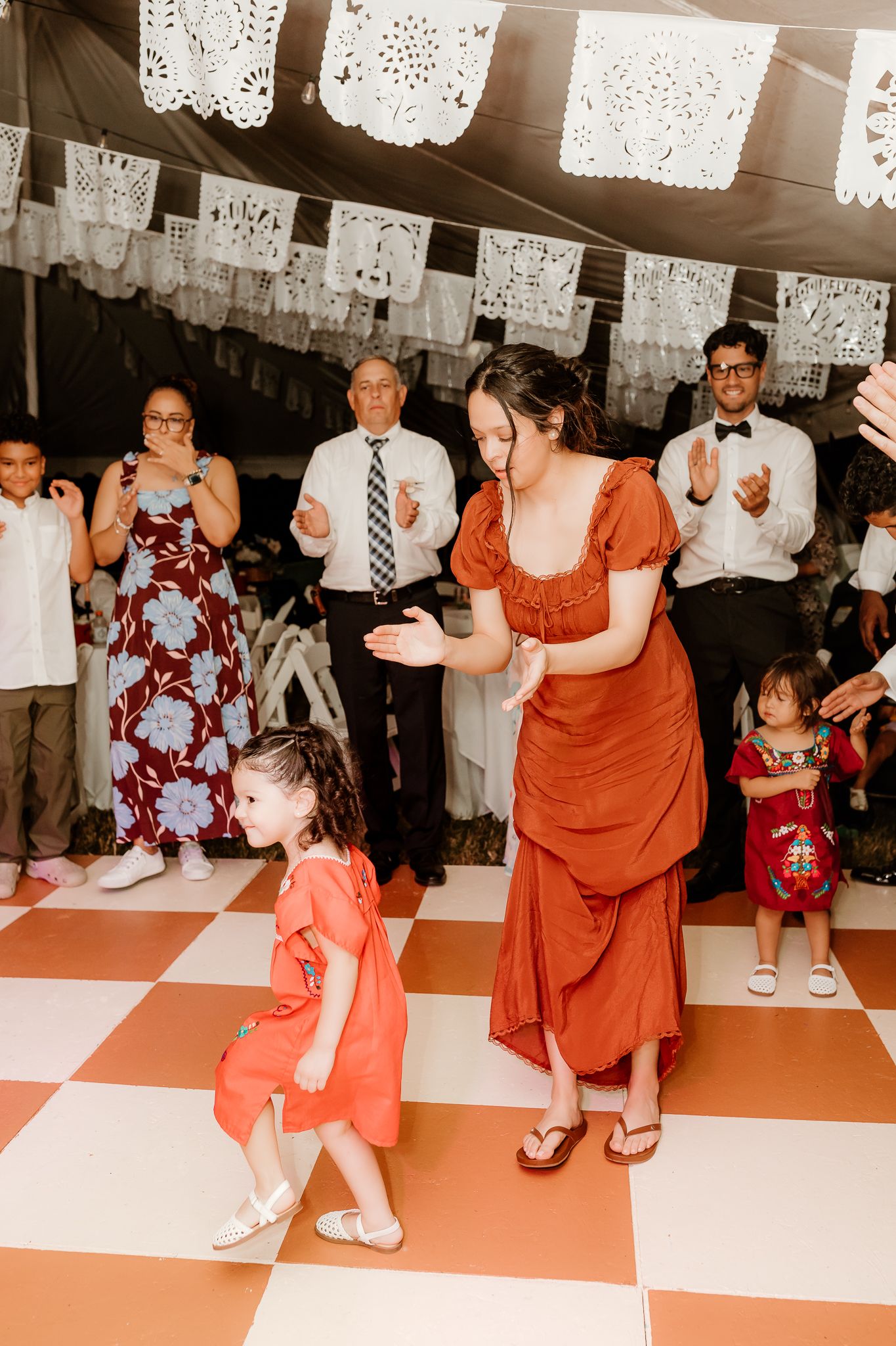 Woman in rust dress dances with little girl in matching outfit at festive celebration with papel picado decorations.