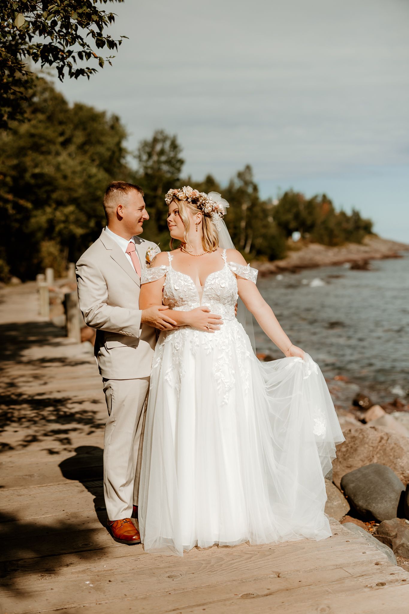 Bride in white floral gown and flower crown walks with groom in tan suit along a scenic lakeside path.