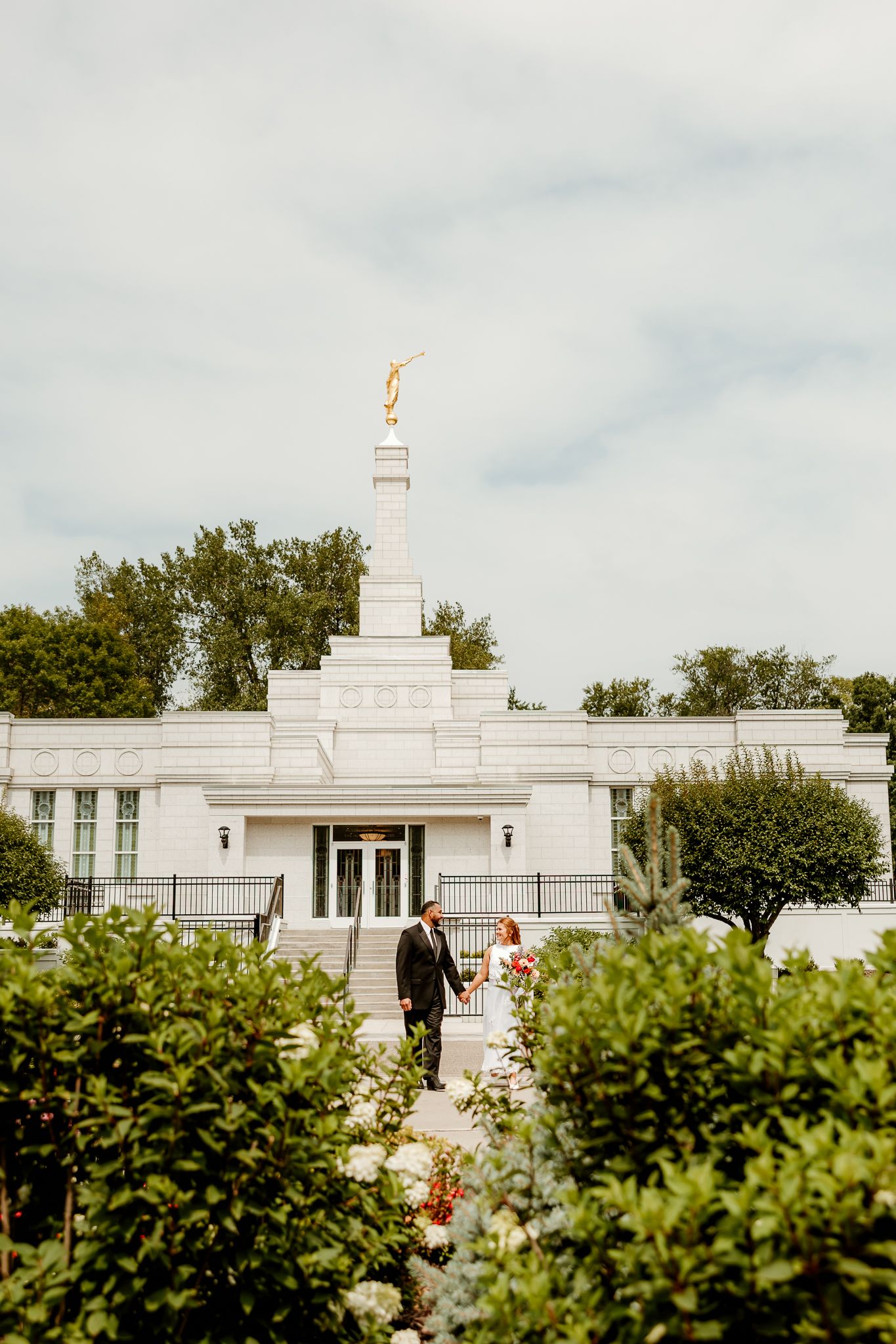 Newlywed couple walking through garden path outside white LDS temple with golden angel Moroni spire.