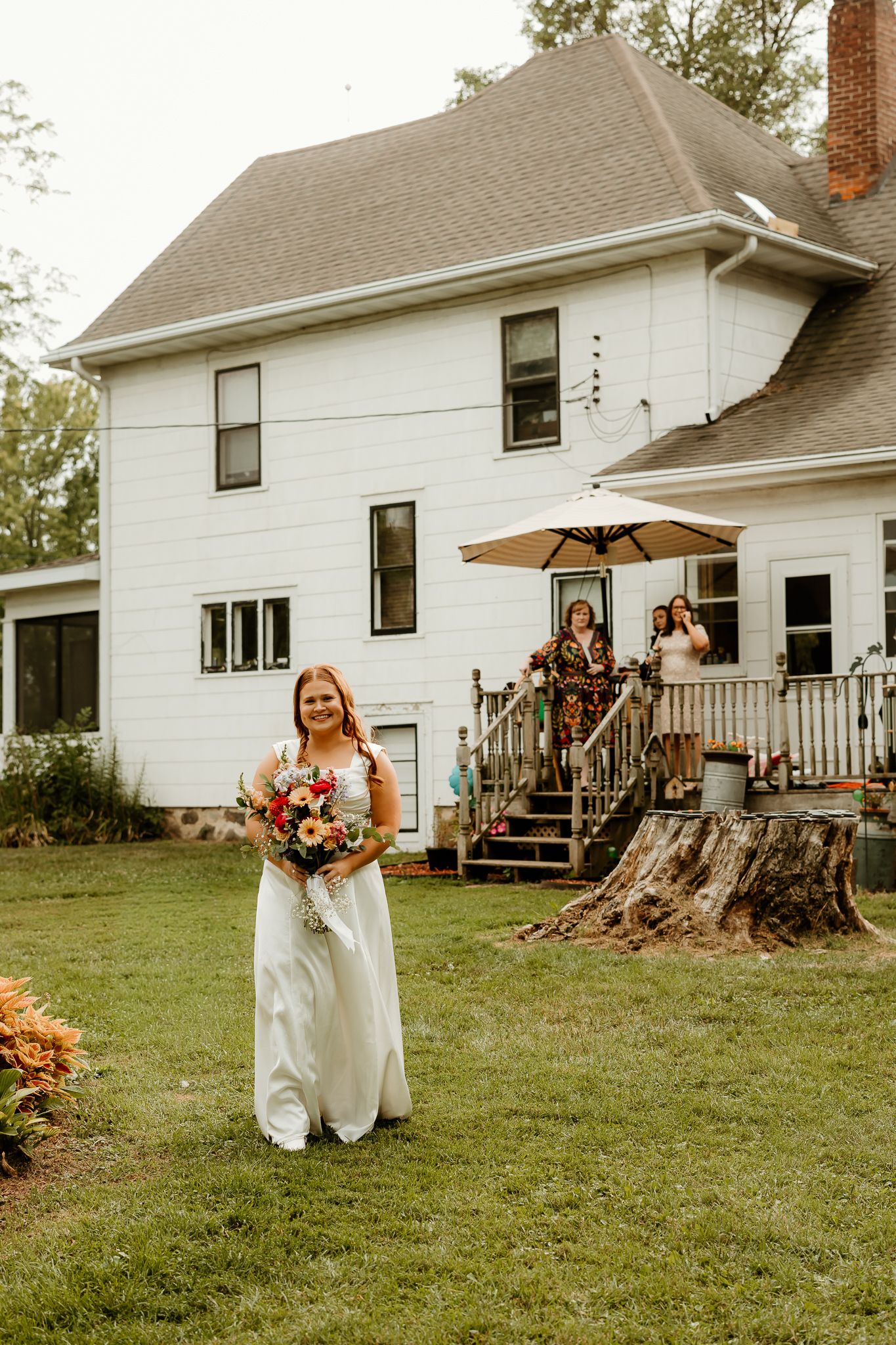 Bride in white gown holding colorful bouquet stands on green lawn outside white farmhouse with guests on porch behind her.