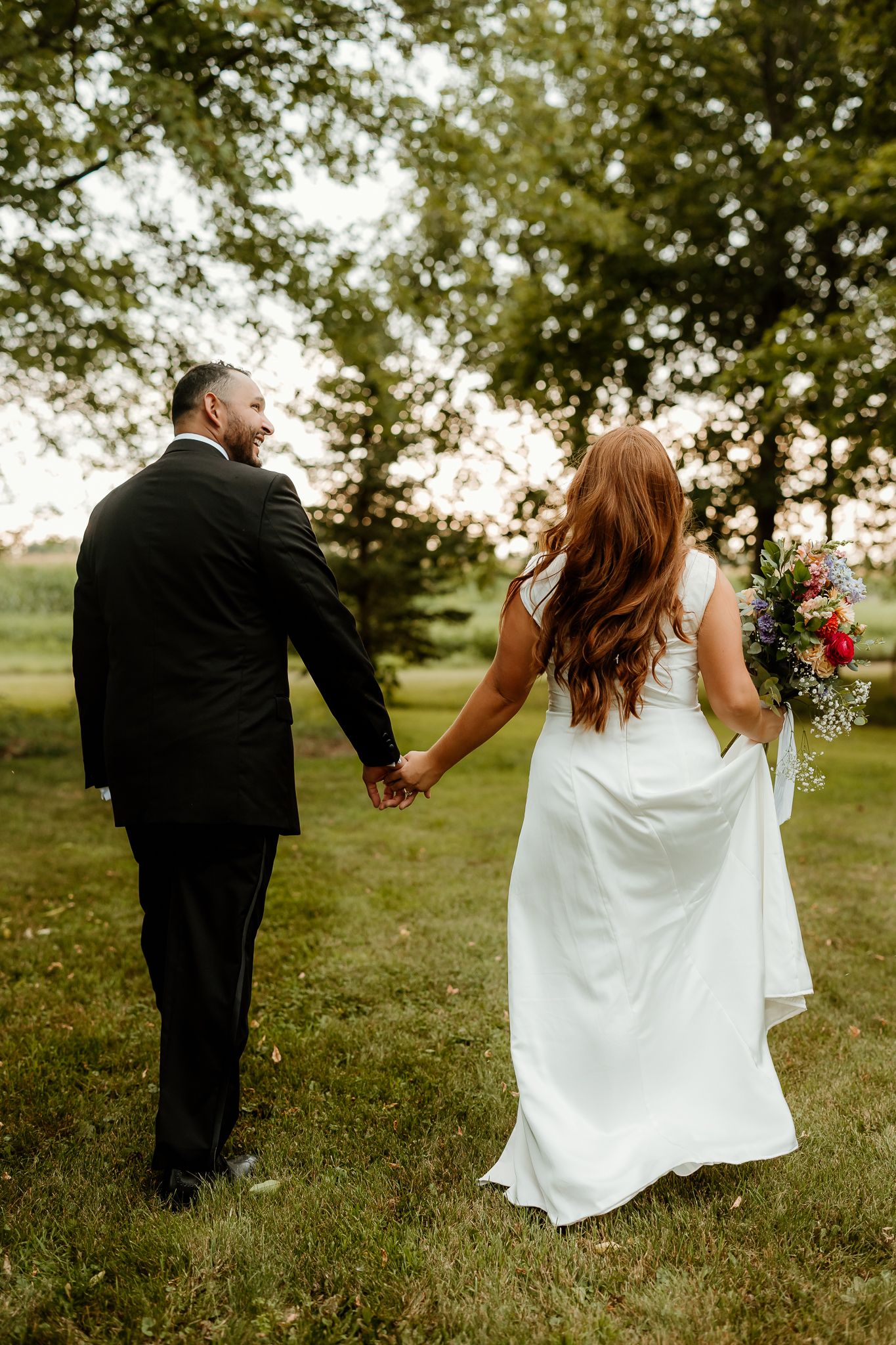 A bride and groom walk hand-in-hand through a lush green garden, she in a white gown holding a colorful bouquet.