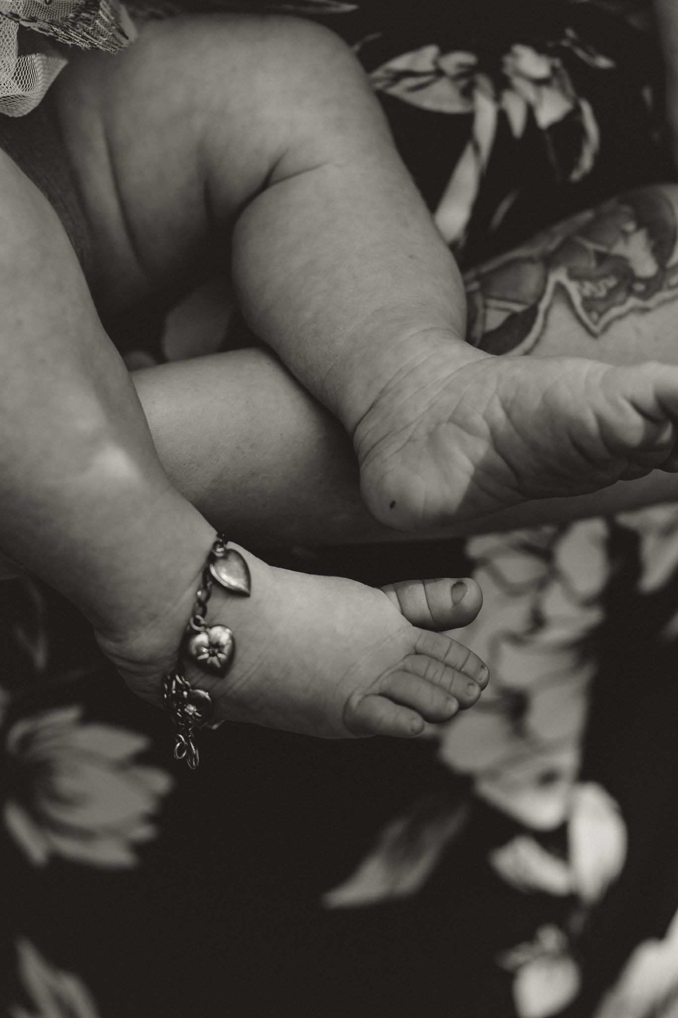 Black and white close-up of a baby's tiny feet and chubby legs, with a delicate charm bracelet on one ankle.