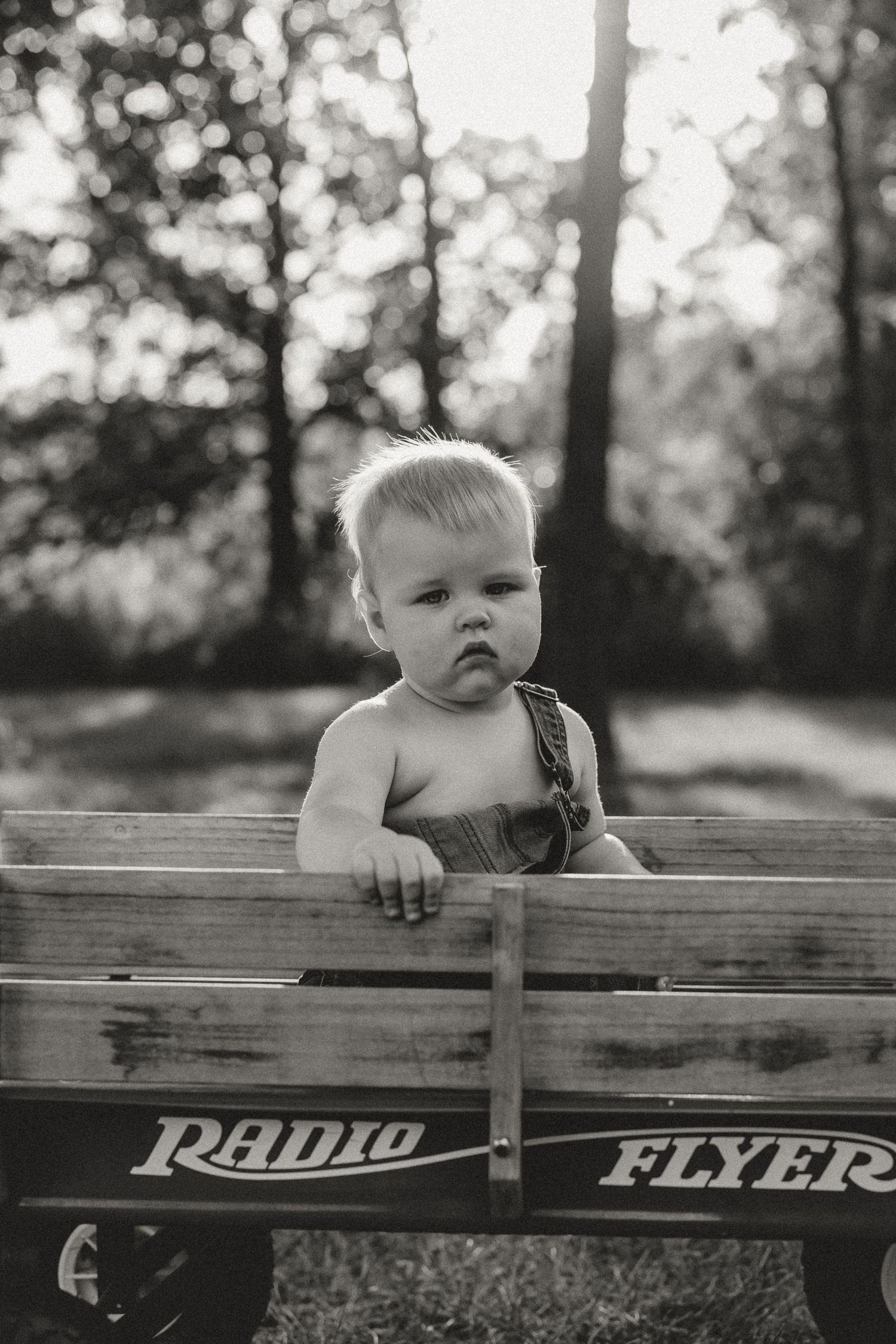 Black and white photo of a baby sitting in a classic Radio Flyer wagon outdoors.