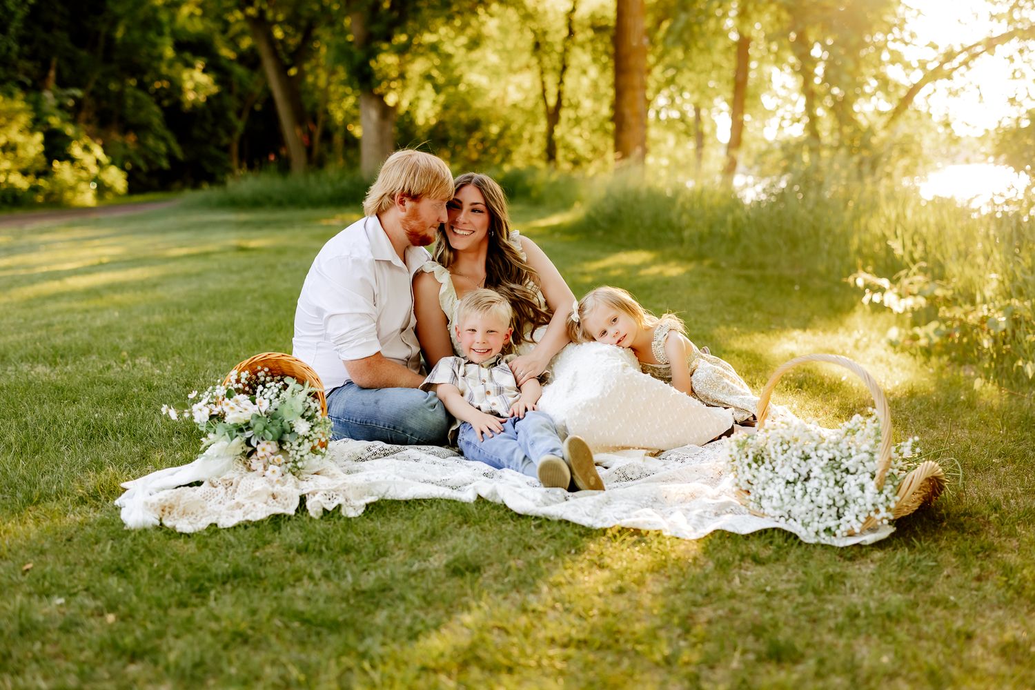 Family of four enjoying a sunny outdoor picnic on a lace blanket surrounded by lush green trees.