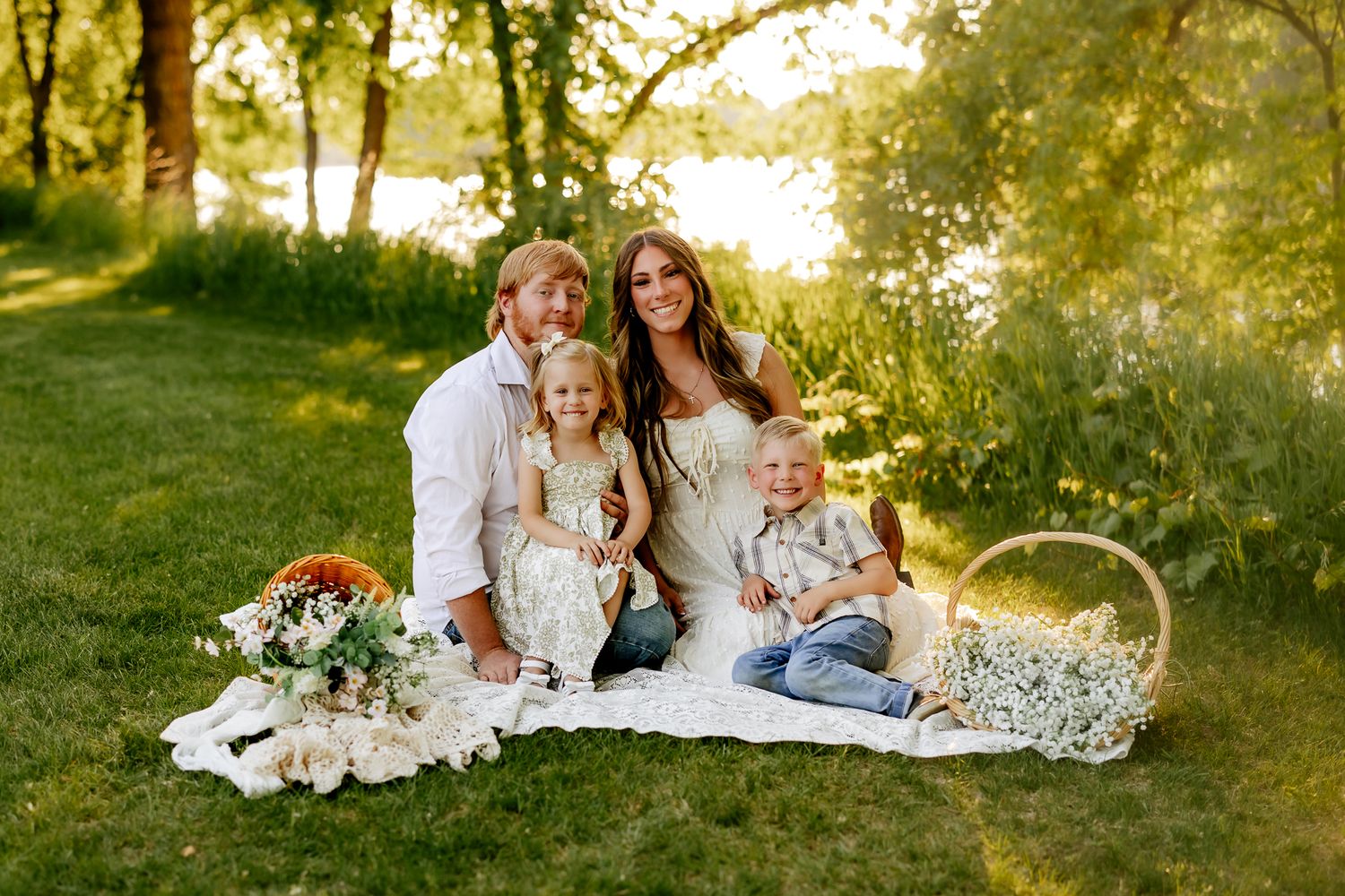Happy family of four sits on white blanket in sunny park with flower baskets nearby.