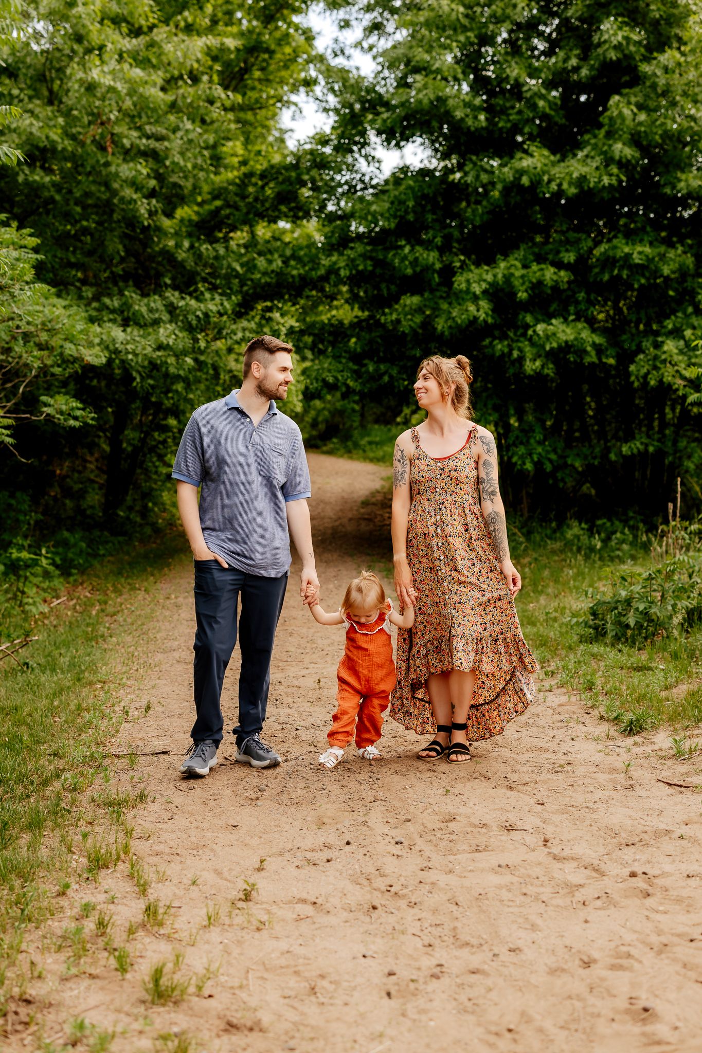 A happy family of three walks hand-in-hand along a tree-lined dirt path during an outdoor photo session.