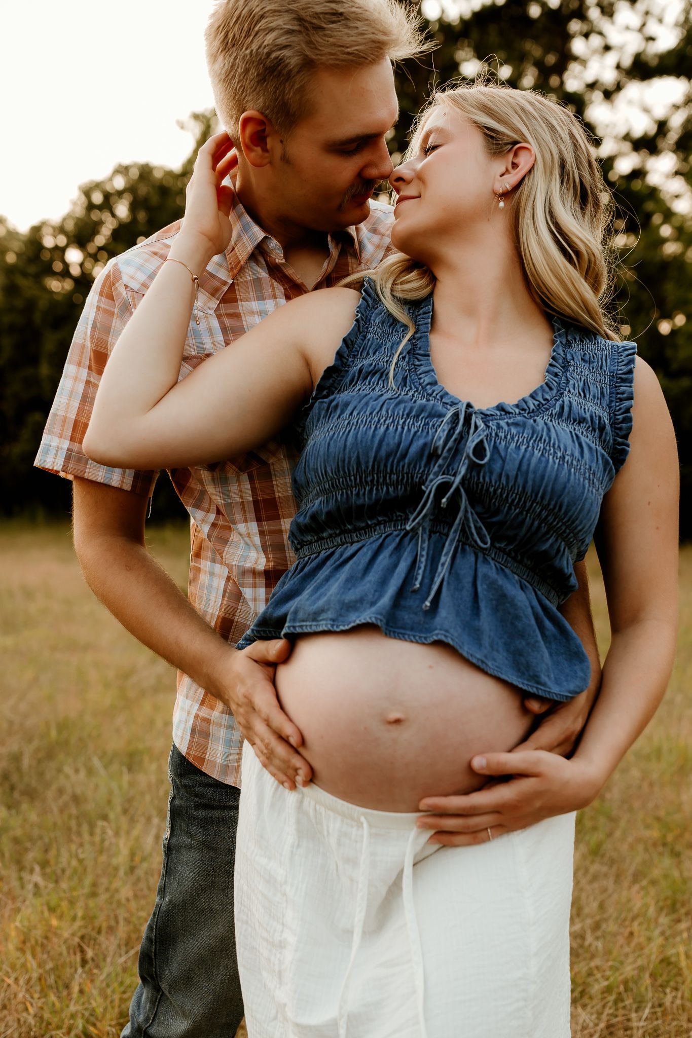 Couple sharing a tender moment outdoors, with pregnant woman's bare belly visible, wearing denim top and white skirt.
