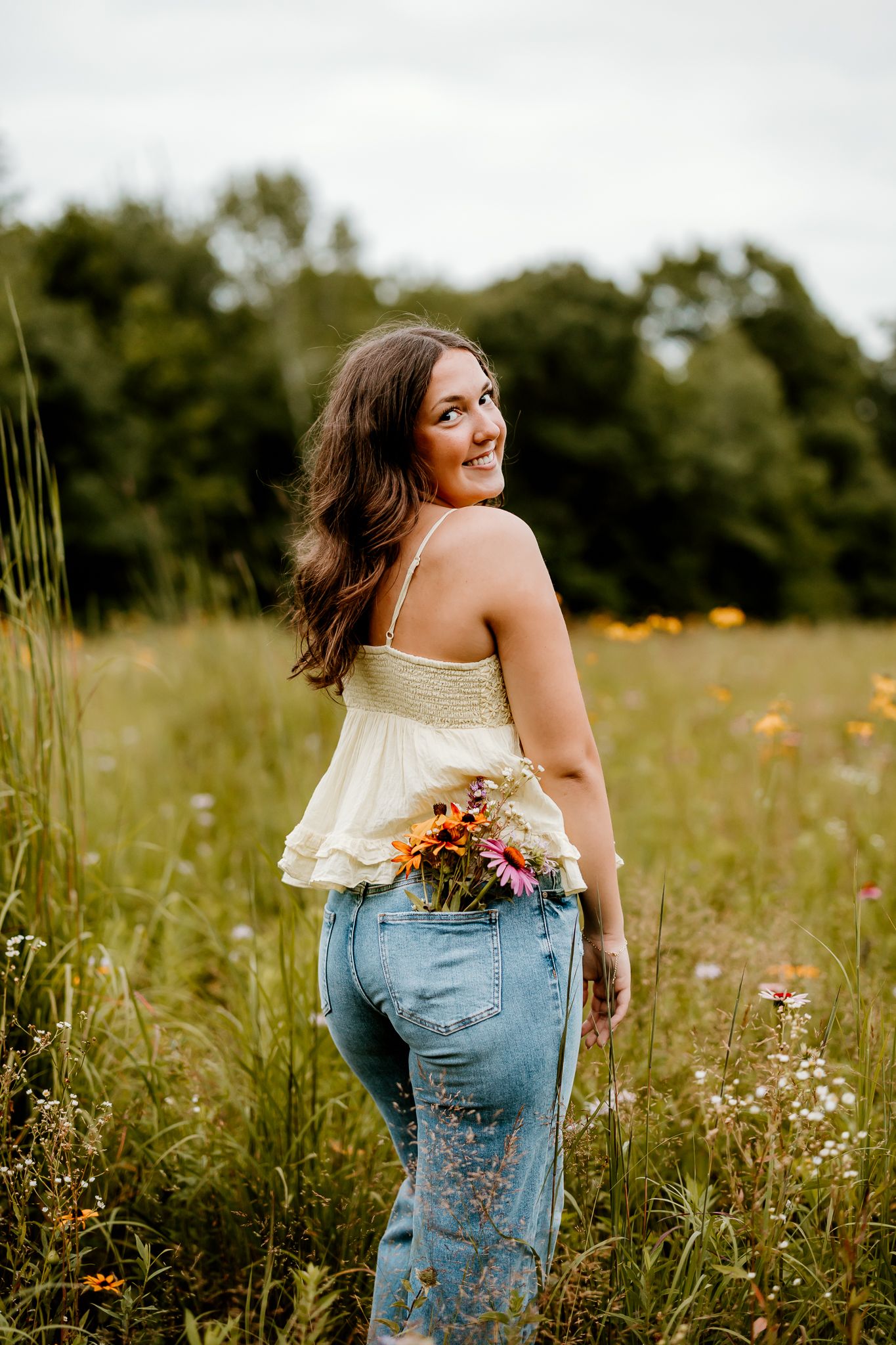 Young woman in white top and jeans holds wildflowers in a meadow, smiling over her shoulder.
