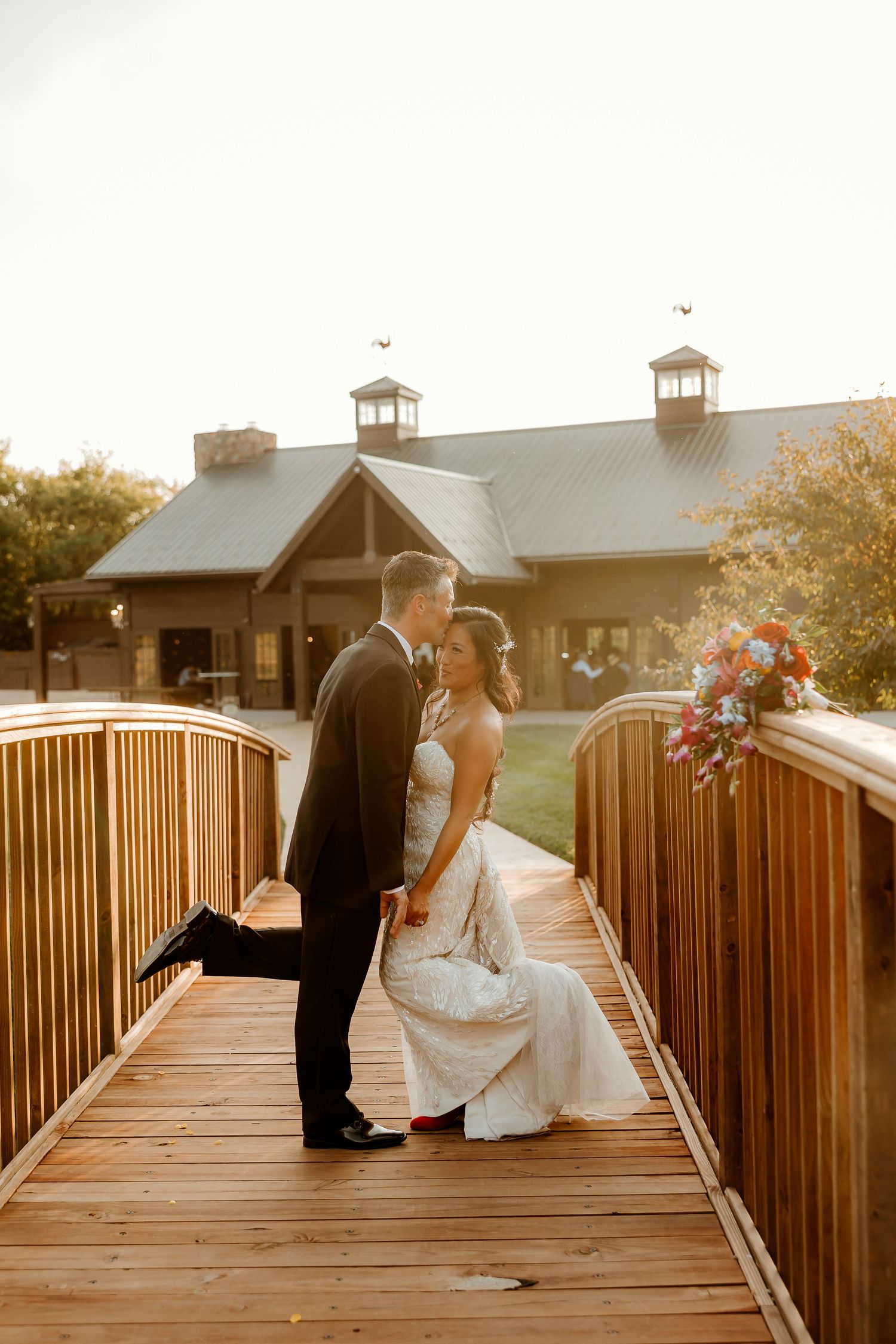 Bride and groom share a romantic moment on a wooden bridge at golden hour outside a rustic wedding venue.