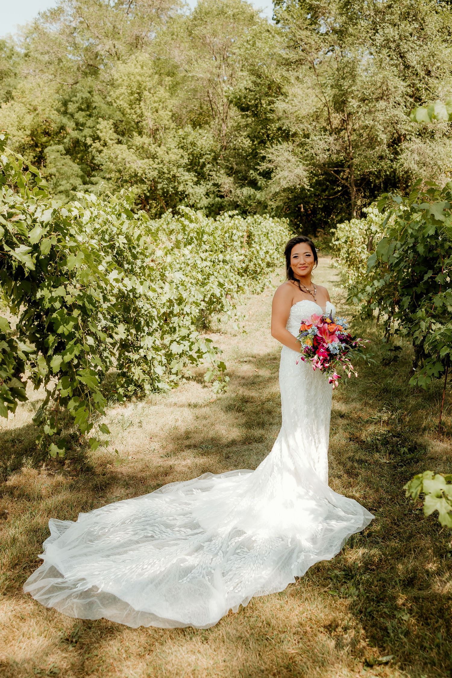 Bride in white mermaid gown with long train holds colorful bouquet among lush vineyard greenery on sunny wedding day.