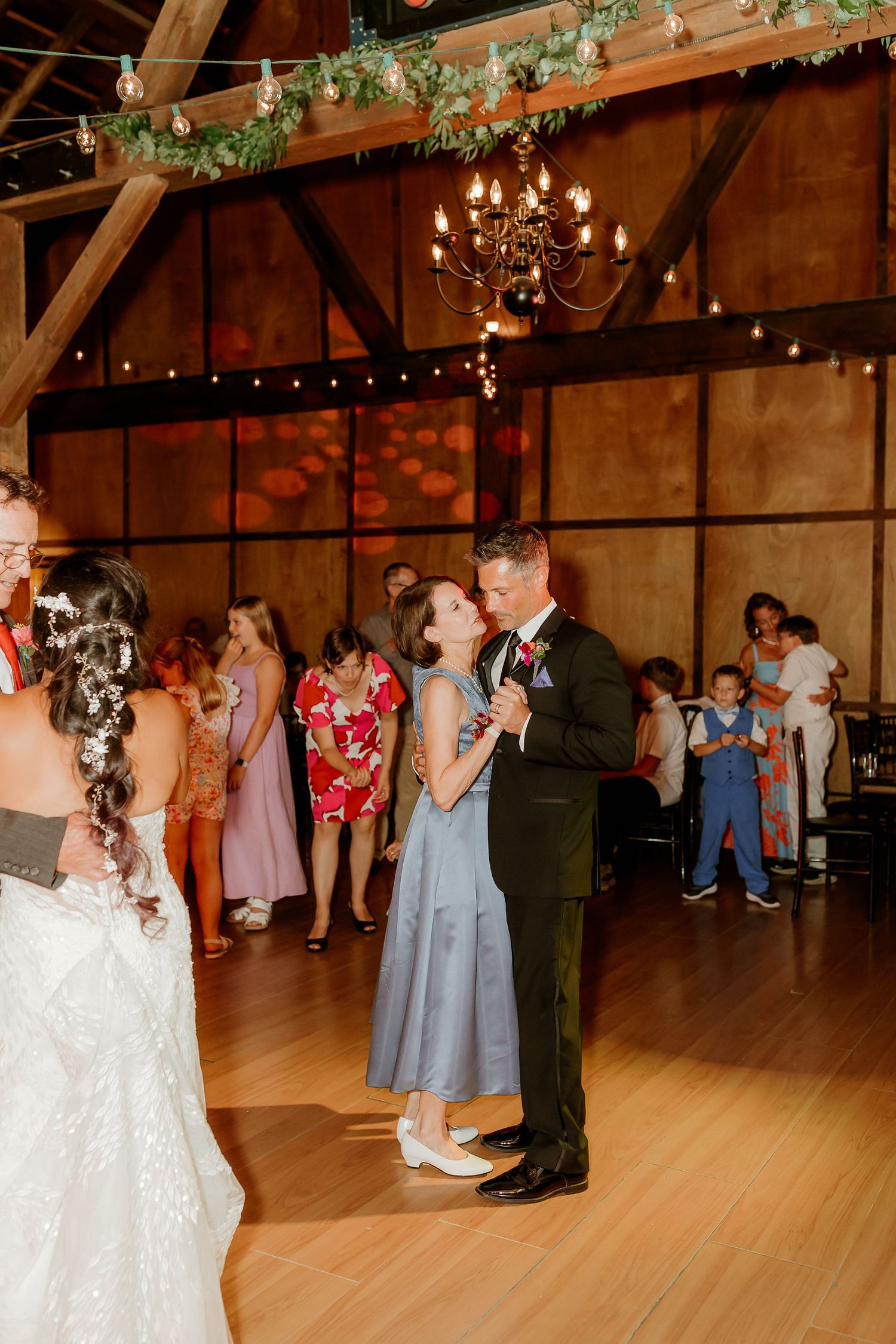 Couple shares a romantic dance at a rustic barn wedding reception with guests watching and string lights overhead.