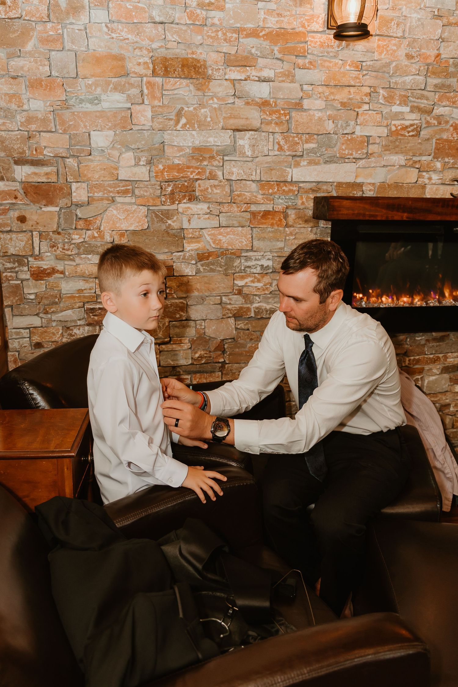A man in a dress shirt and tie helps a young boy with his outfit while seated in leather chairs near a stone fireplace.