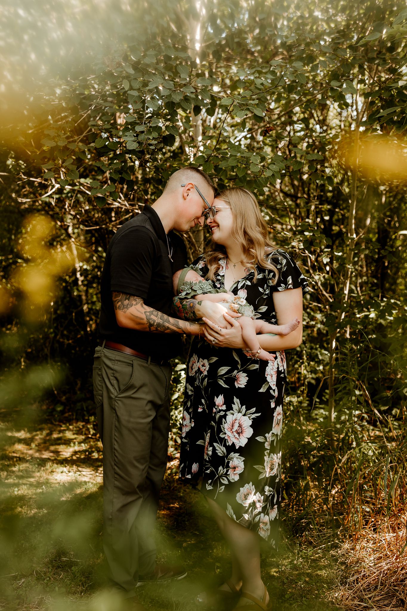 A couple shares a tender moment holding their newborn baby outdoors, surrounded by lush green foliage in warm golden light.