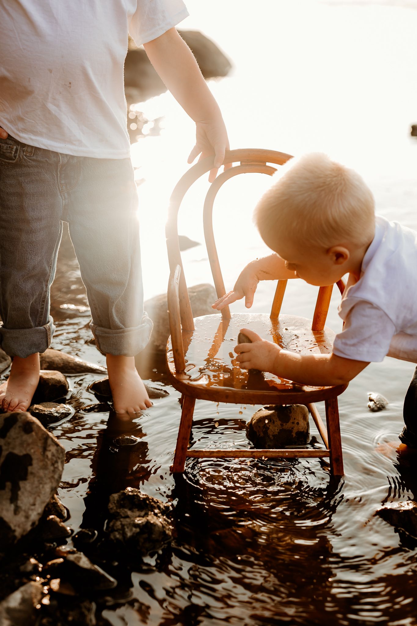 A toddler and adult play with a wooden chair in shallow water during golden hour sunset light.