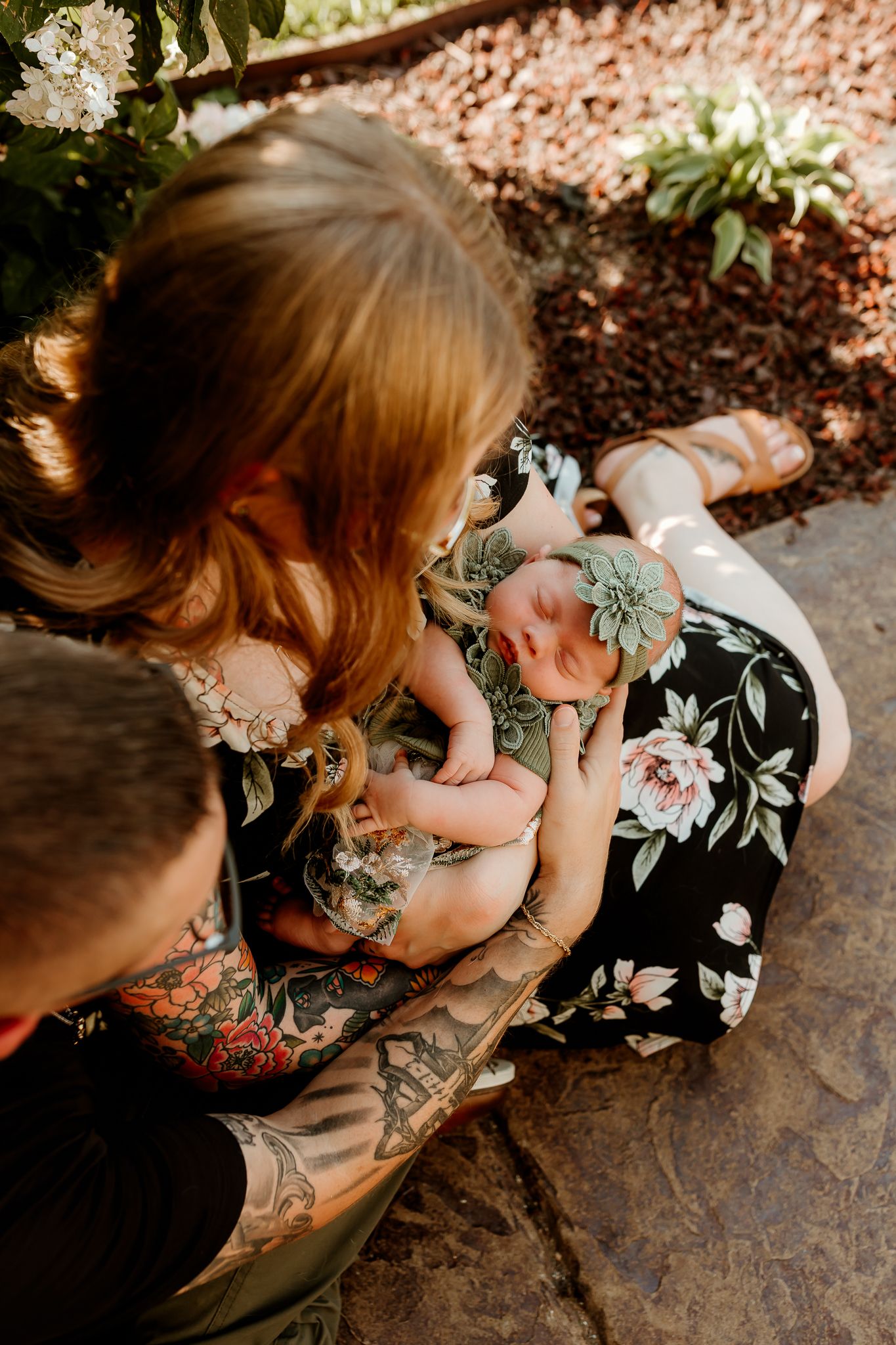New parents with tattoos lovingly cradle their sleeping newborn baby wearing a floral outfit and green bow headband.