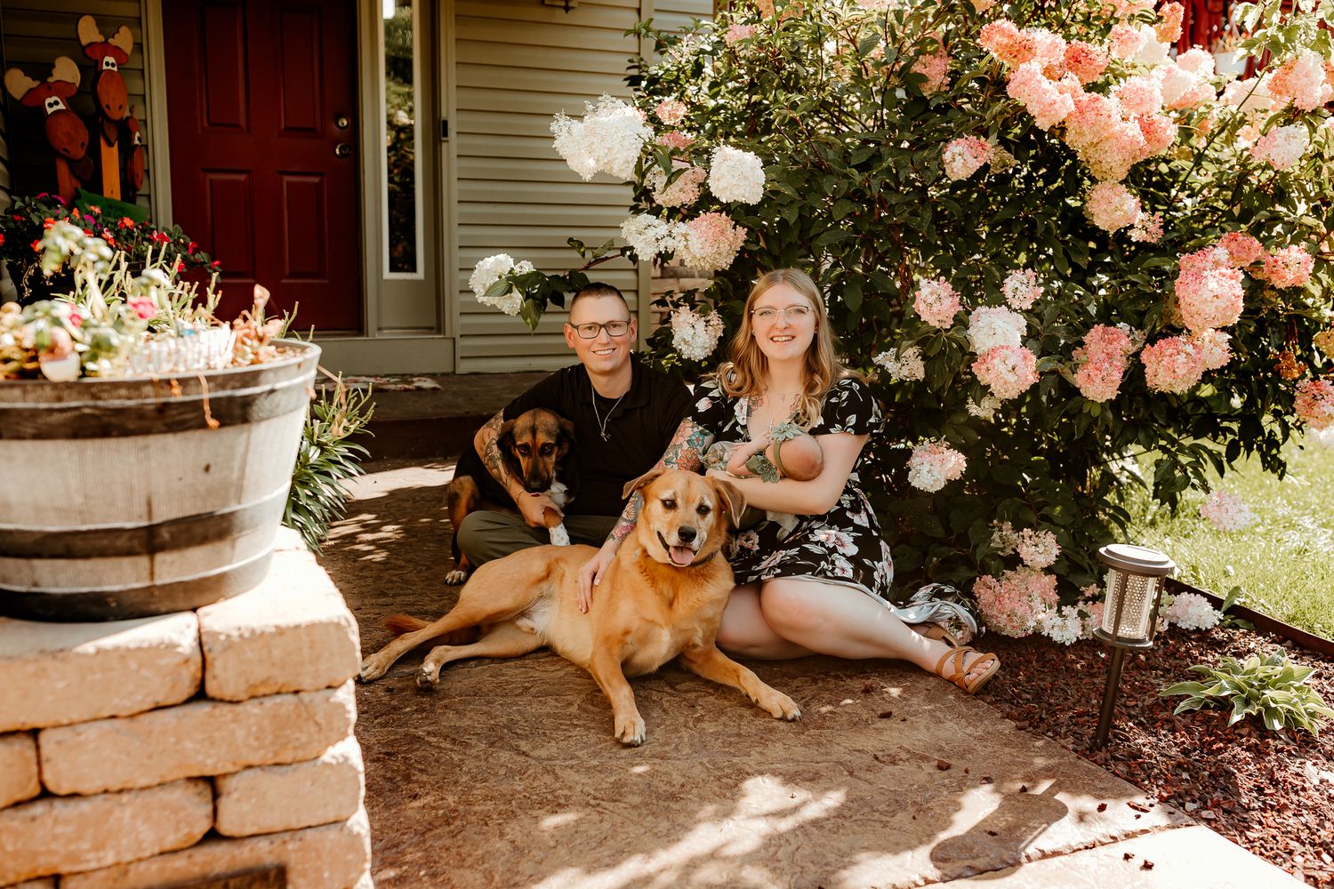 Couple sitting with two dogs in front of a flowering rose bush outside a charming home entrance.