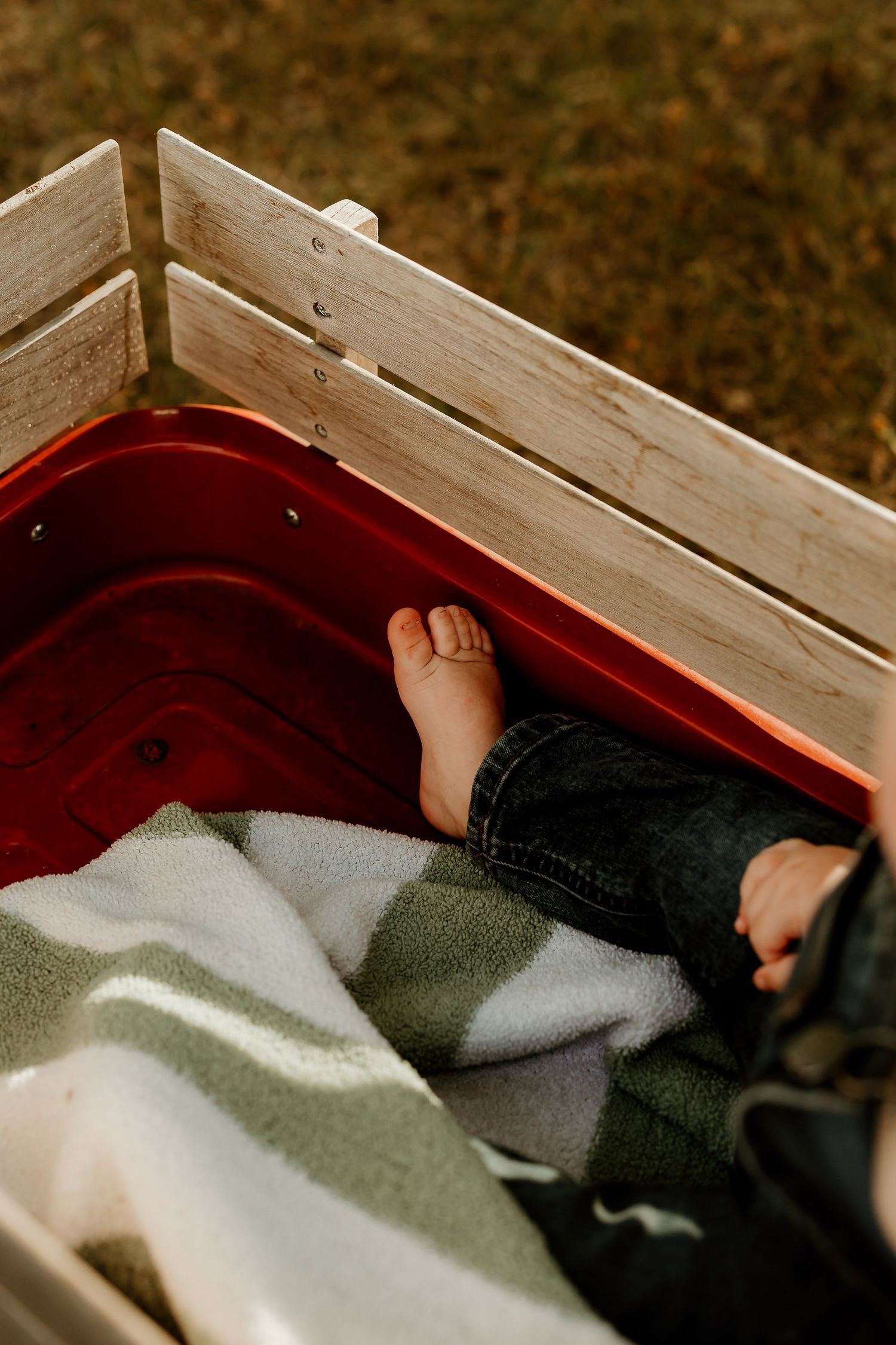 Bare toddler feet peeking out from a cozy plaid blanket inside a classic red wooden wagon outdoors.