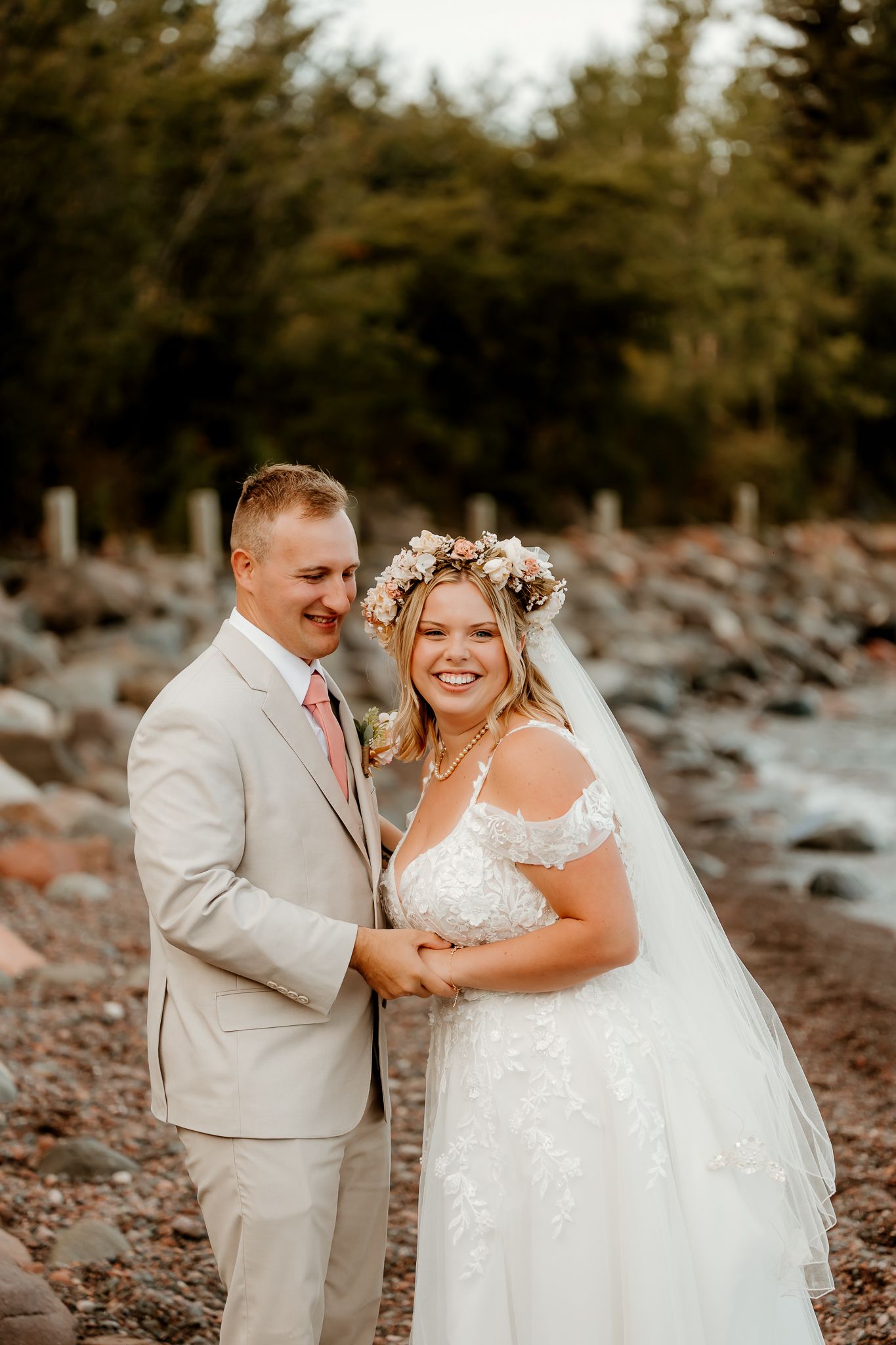 Smiling bride in floral crown and groom in tan suit pose together on rocky shoreline with trees behind.