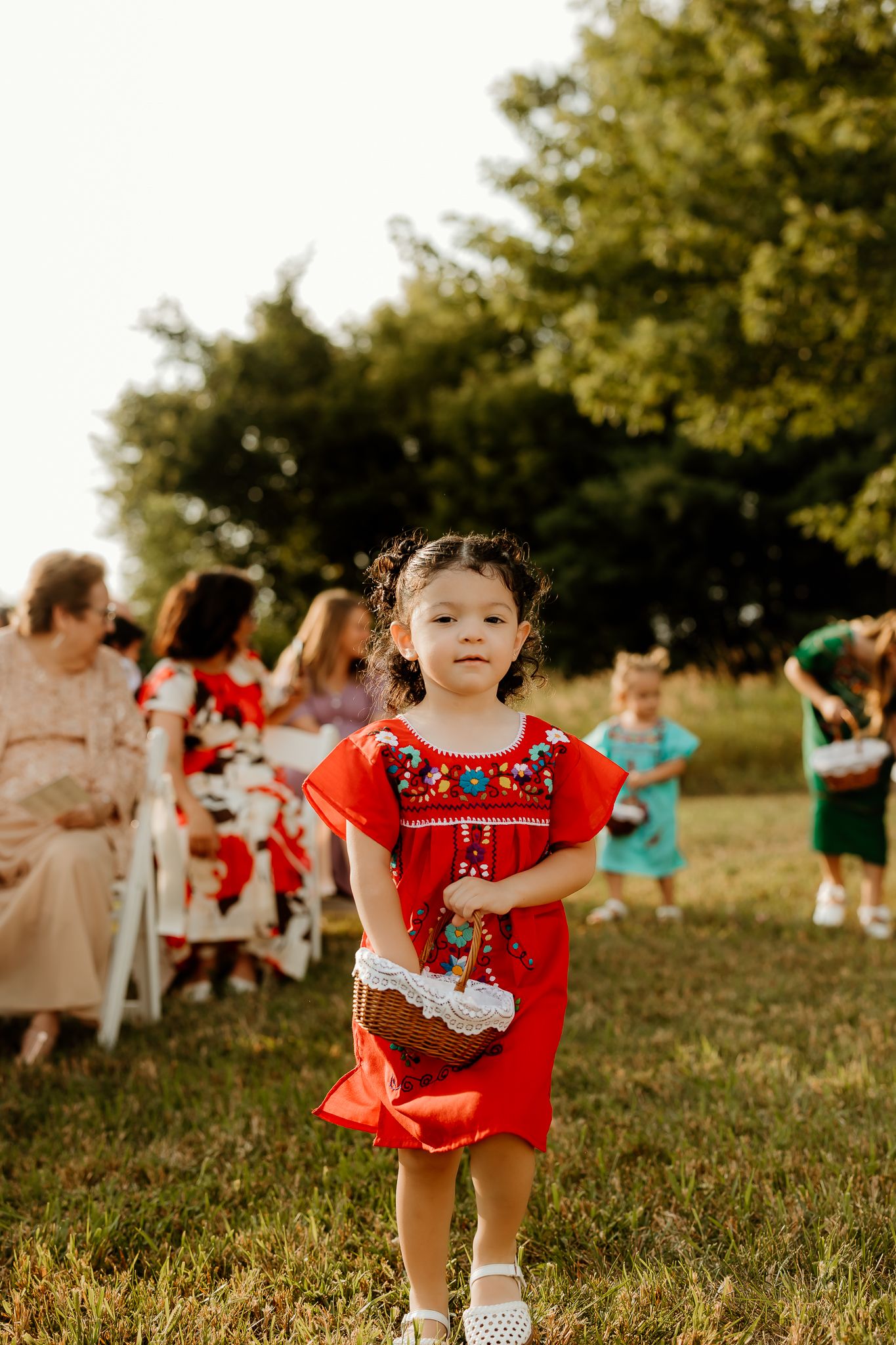A young girl in a red embroidered dress walks down an outdoor wedding aisle carrying a heart-shaped basket.