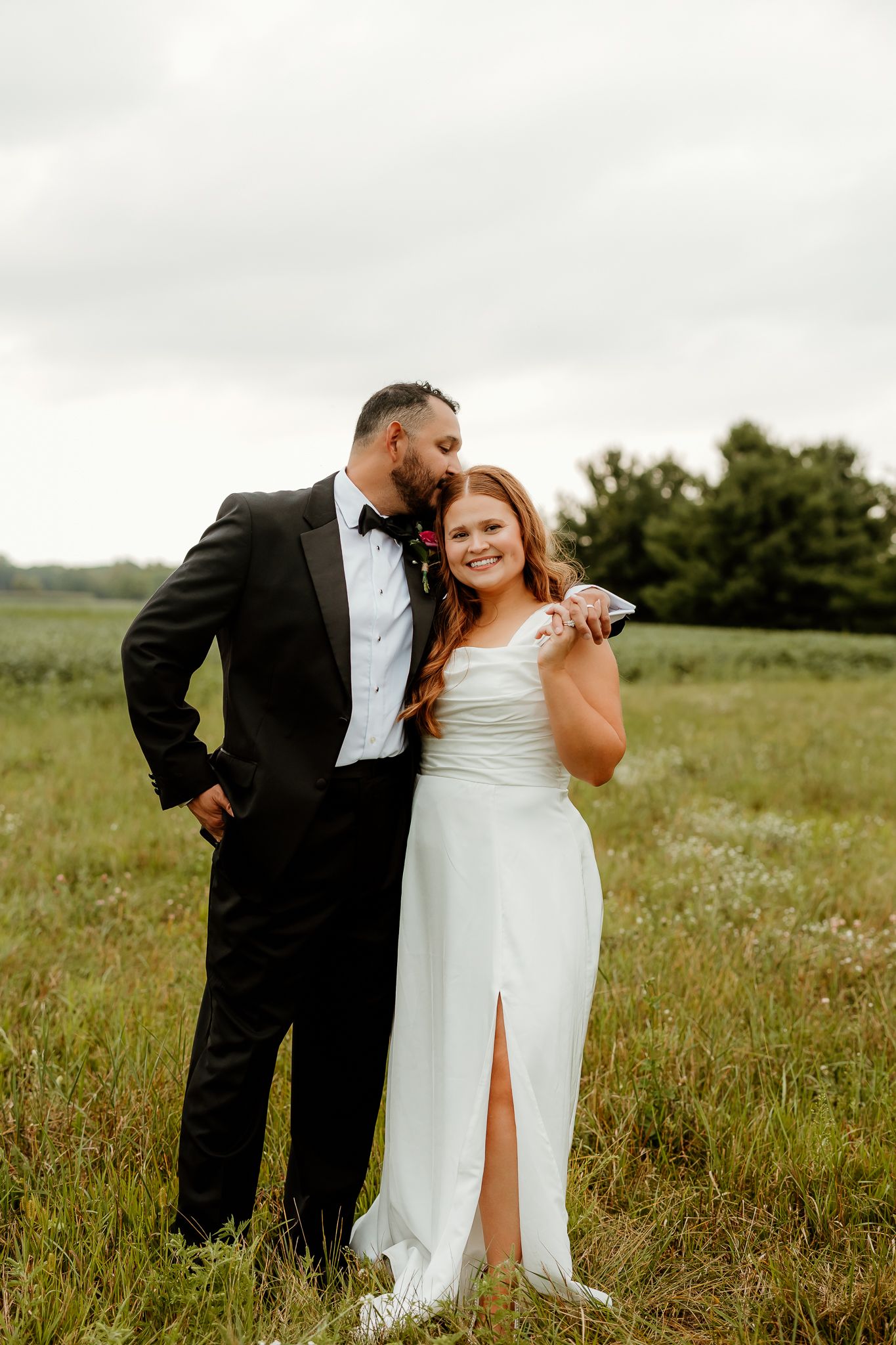 Bride in white gown and groom in black tuxedo pose together in a lush green field on their wedding day.
