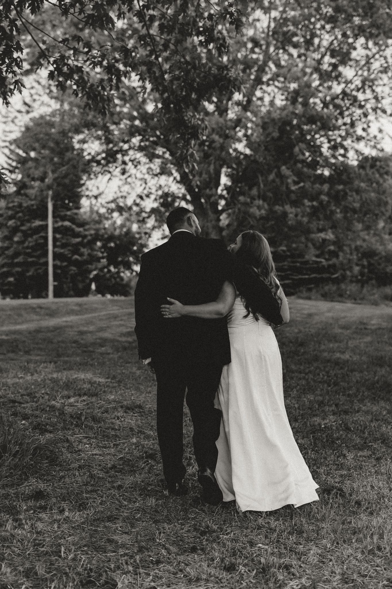 Black and white photo of a bride and groom embracing from behind in a lush green park setting.