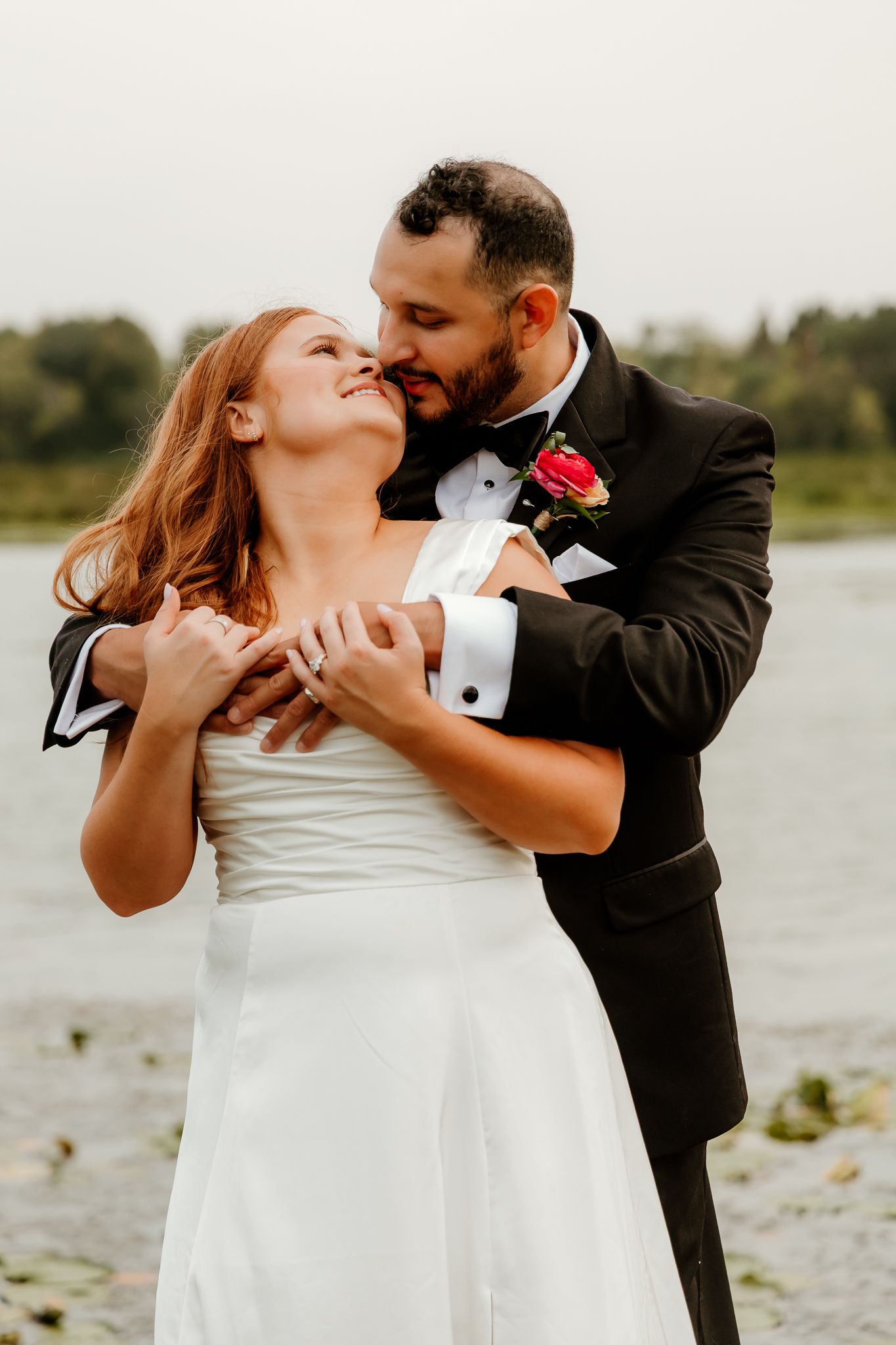 A bride in a white gown and groom in a black tuxedo share a romantic embrace near a serene lakeside backdrop.