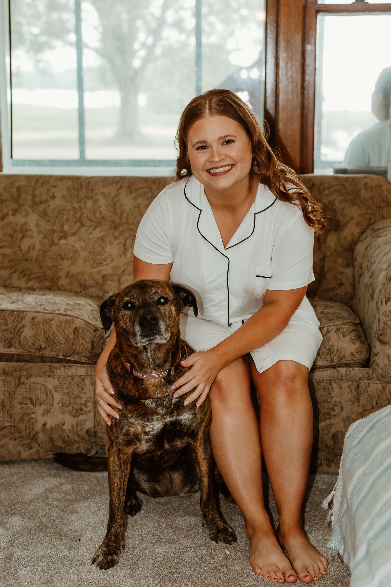 Smiling young woman in white pajamas sitting on a couch with her brindle dog on a carpeted floor.
