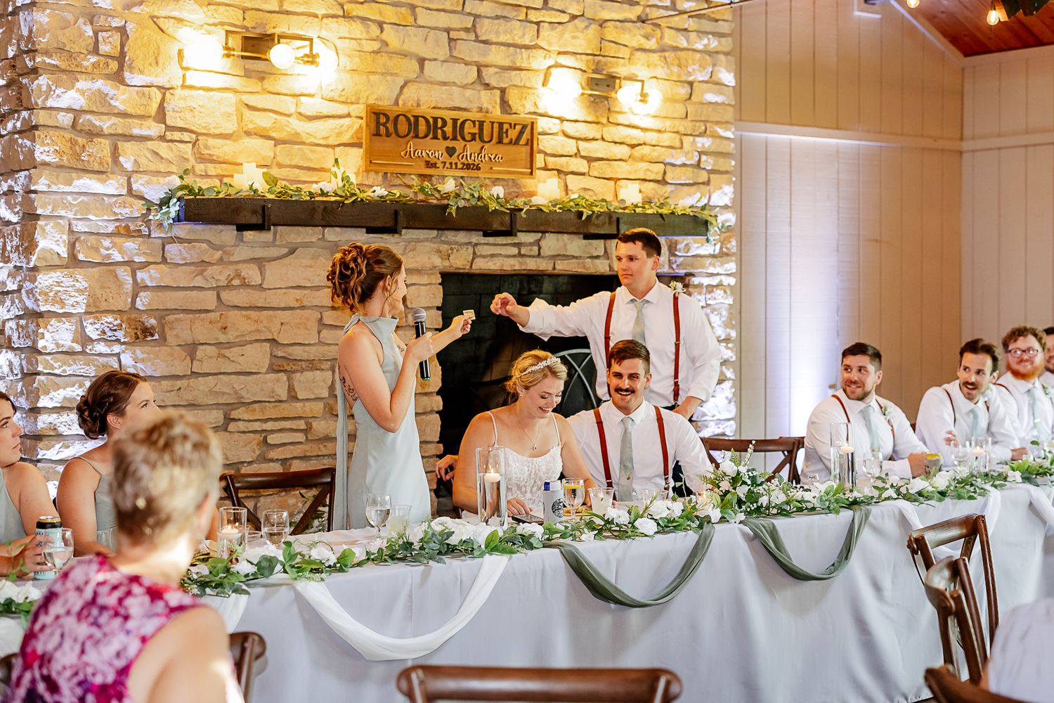 Wedding reception toast at Rodriguez venue with bridal party at decorated head table near stone fireplace.