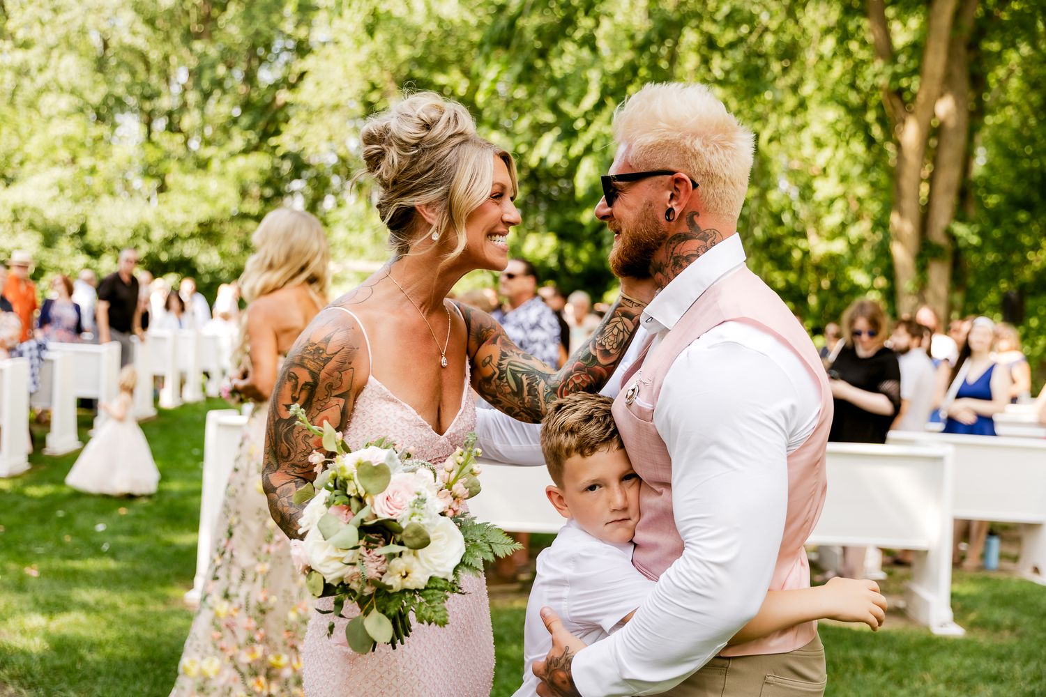 Bride and groom with tattooed arms share a joyful moment with young boy at outdoor wedding ceremony.