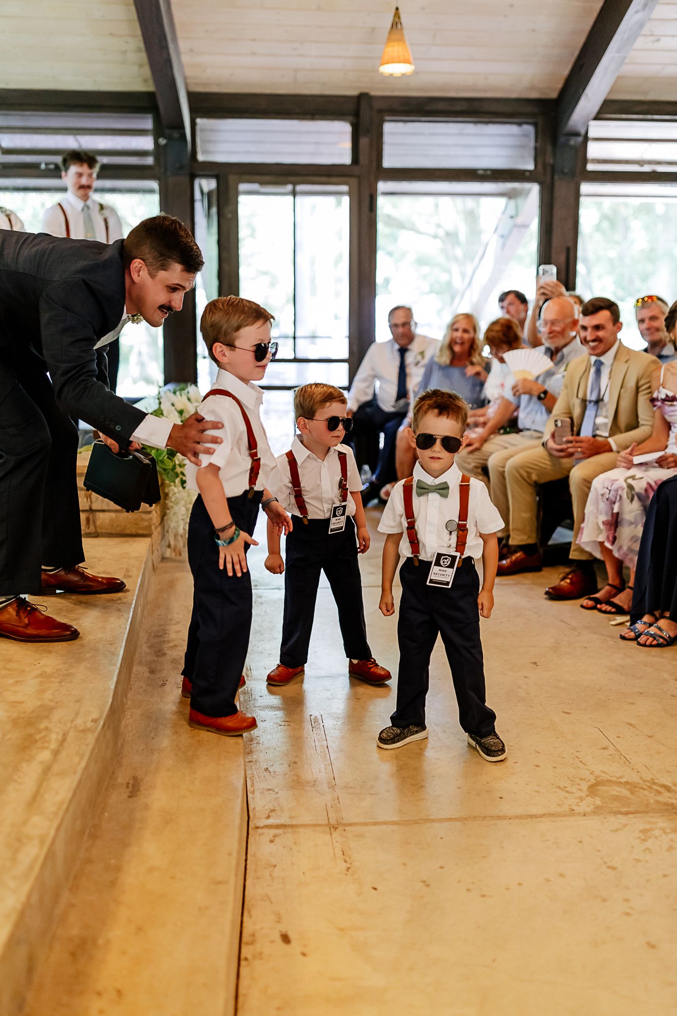 Three young ring bearers in suspenders and sunglasses walk down the aisle at a wedding ceremony.