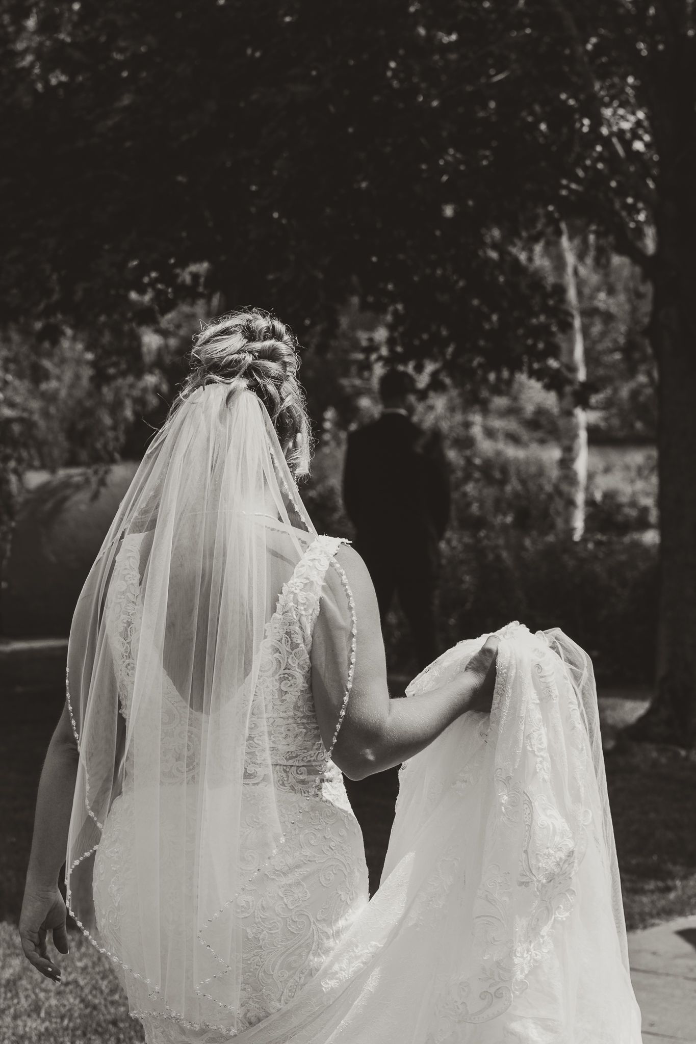 Black and white photo of a bride in a lace wedding dress and veil, seen from behind, walking outdoors.