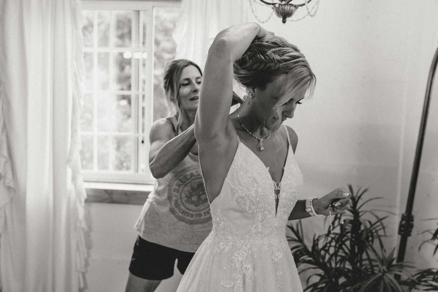 Bride being helped into her lace wedding dress by bridesmaid in black and white photo.
