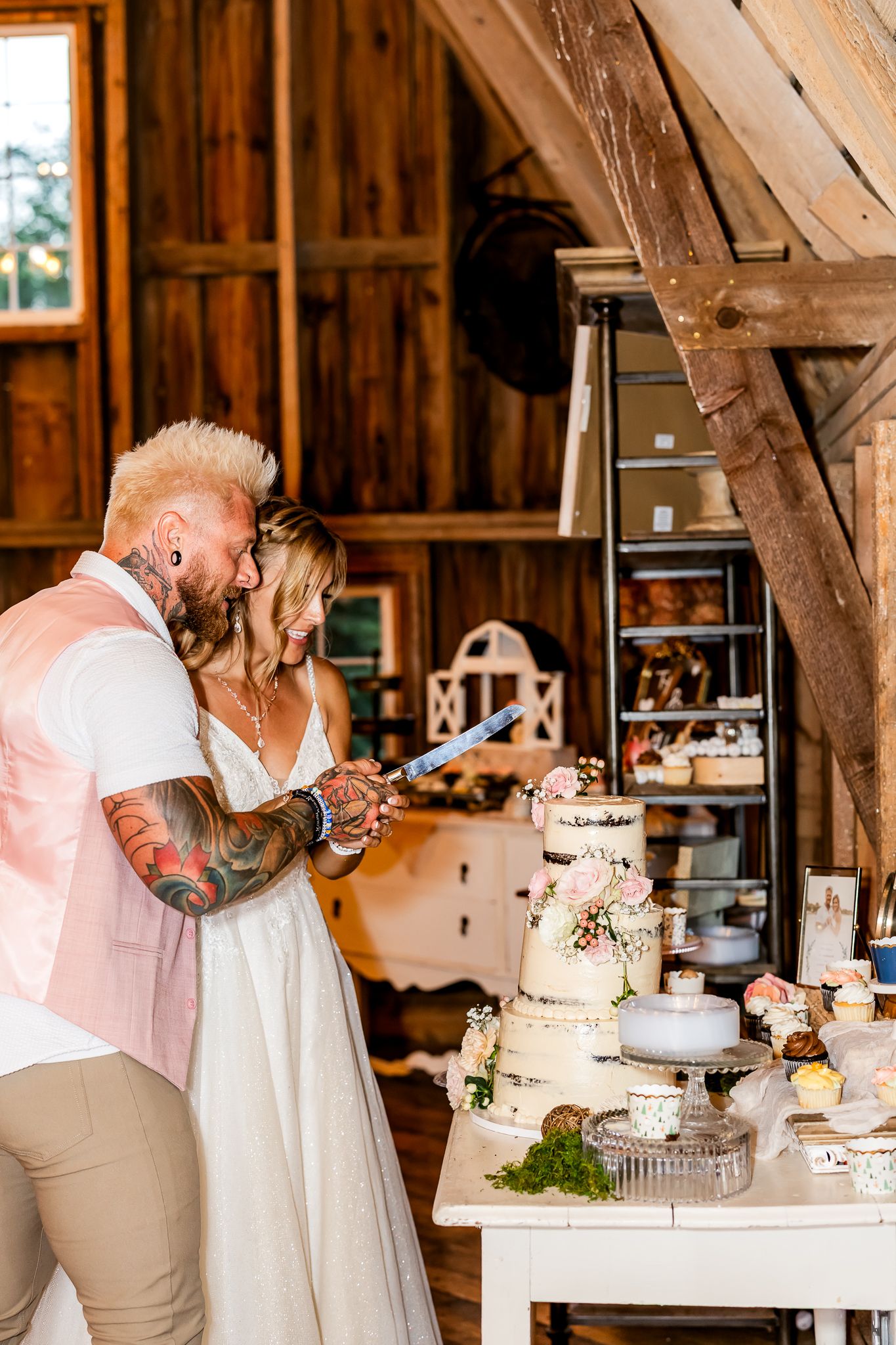 Tattooed groom and bride cutting wedding cake in rustic barn venue.