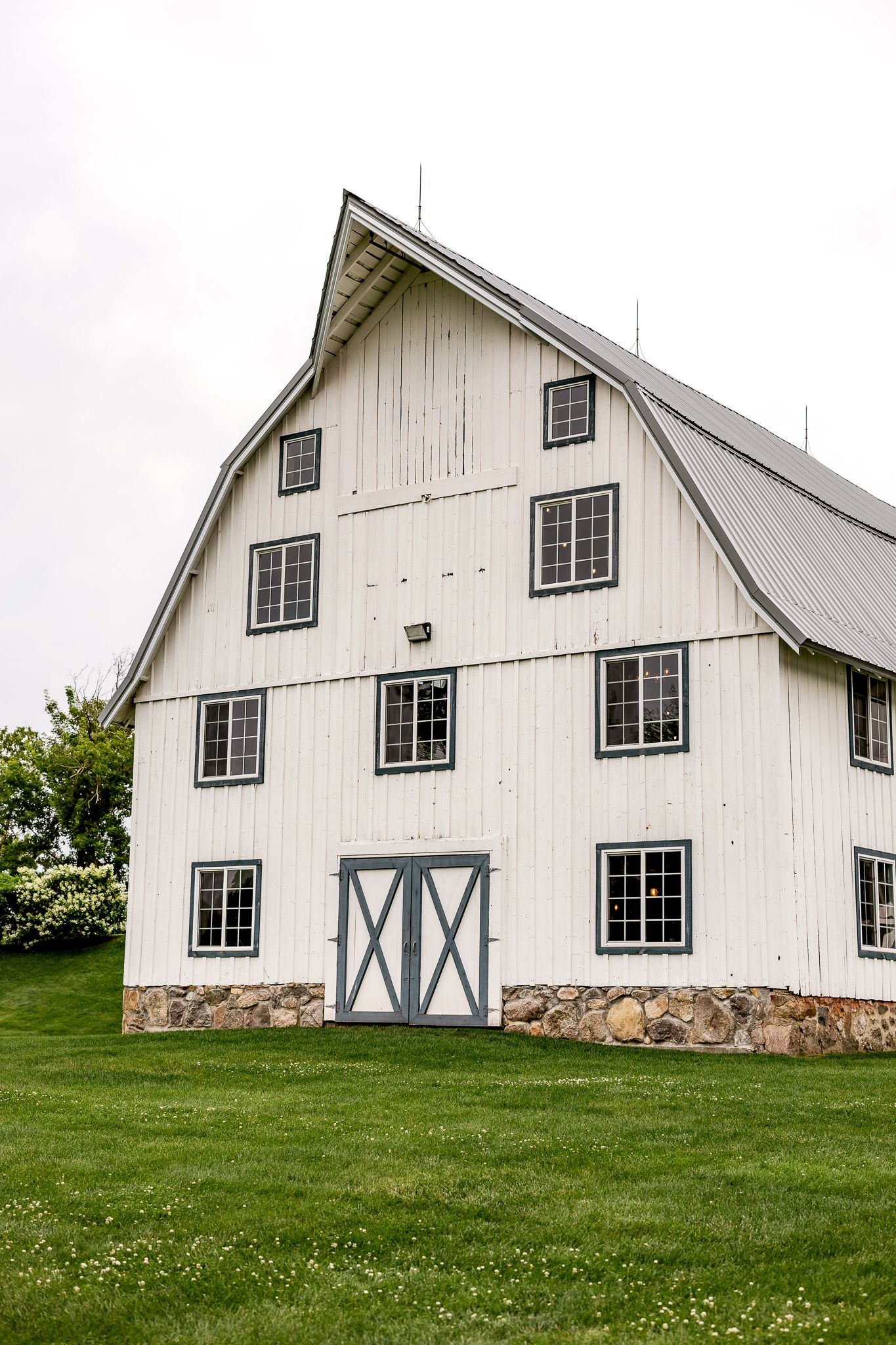 White barn with gray trim, multiple windows, and a gambrel roof sits on a lush green lawn under an overcast sky.