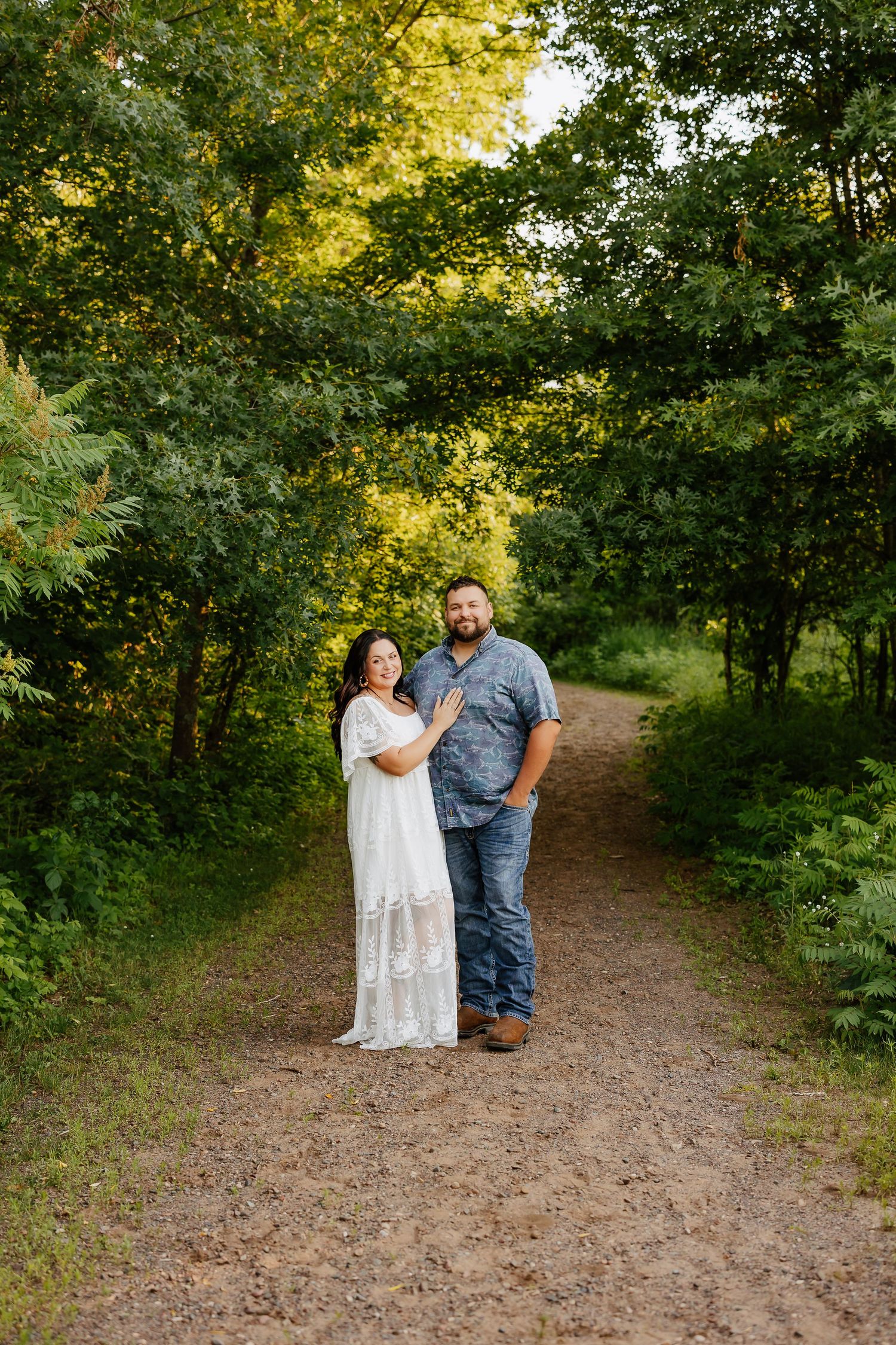 Expecting couple posing on a scenic dirt path surrounded by lush green trees in golden light.