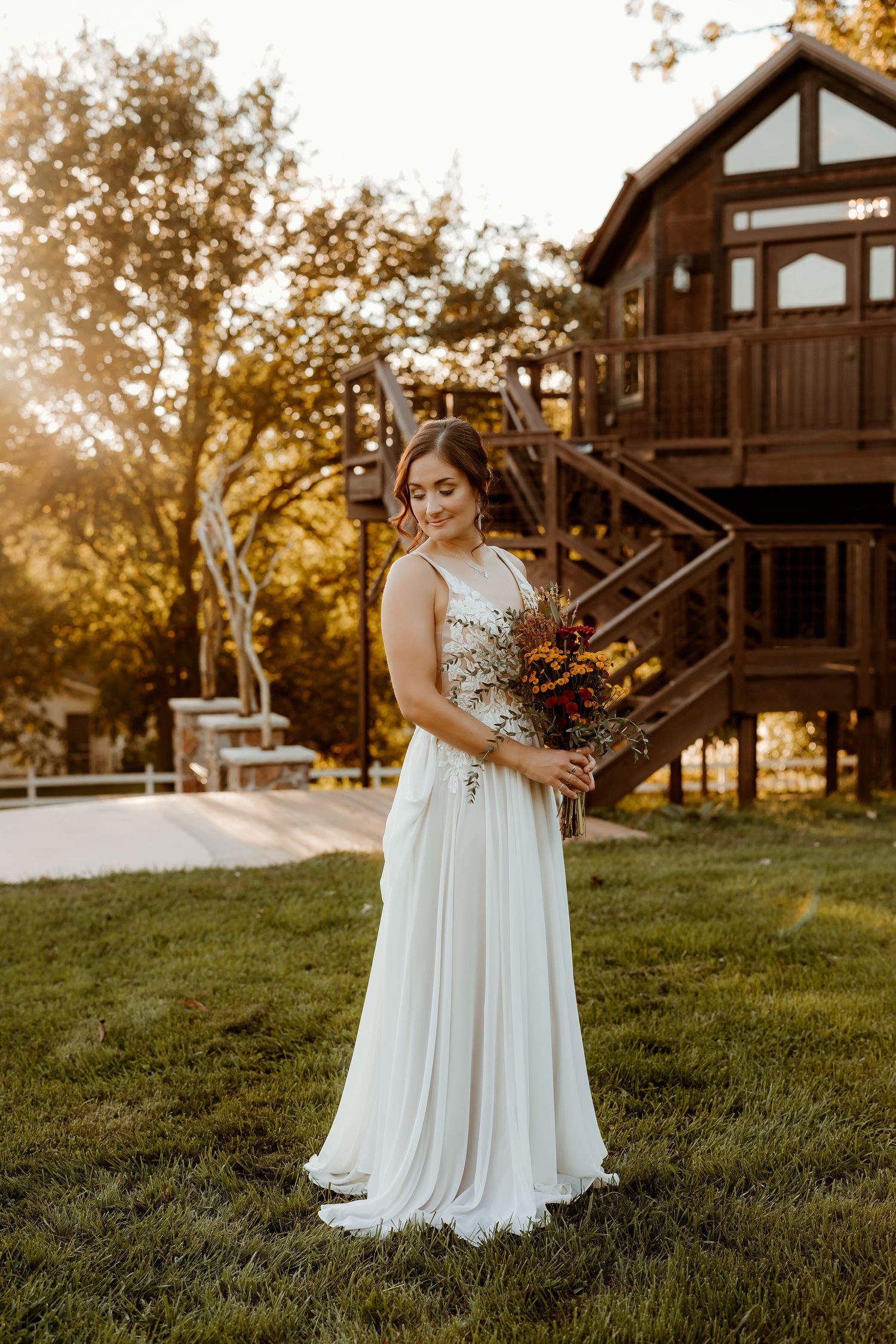 Bride in a flowing white gown with a lace bodice holds an autumn bouquet on a grassy lawn beside a rustic wooden treehouse at golden hour.