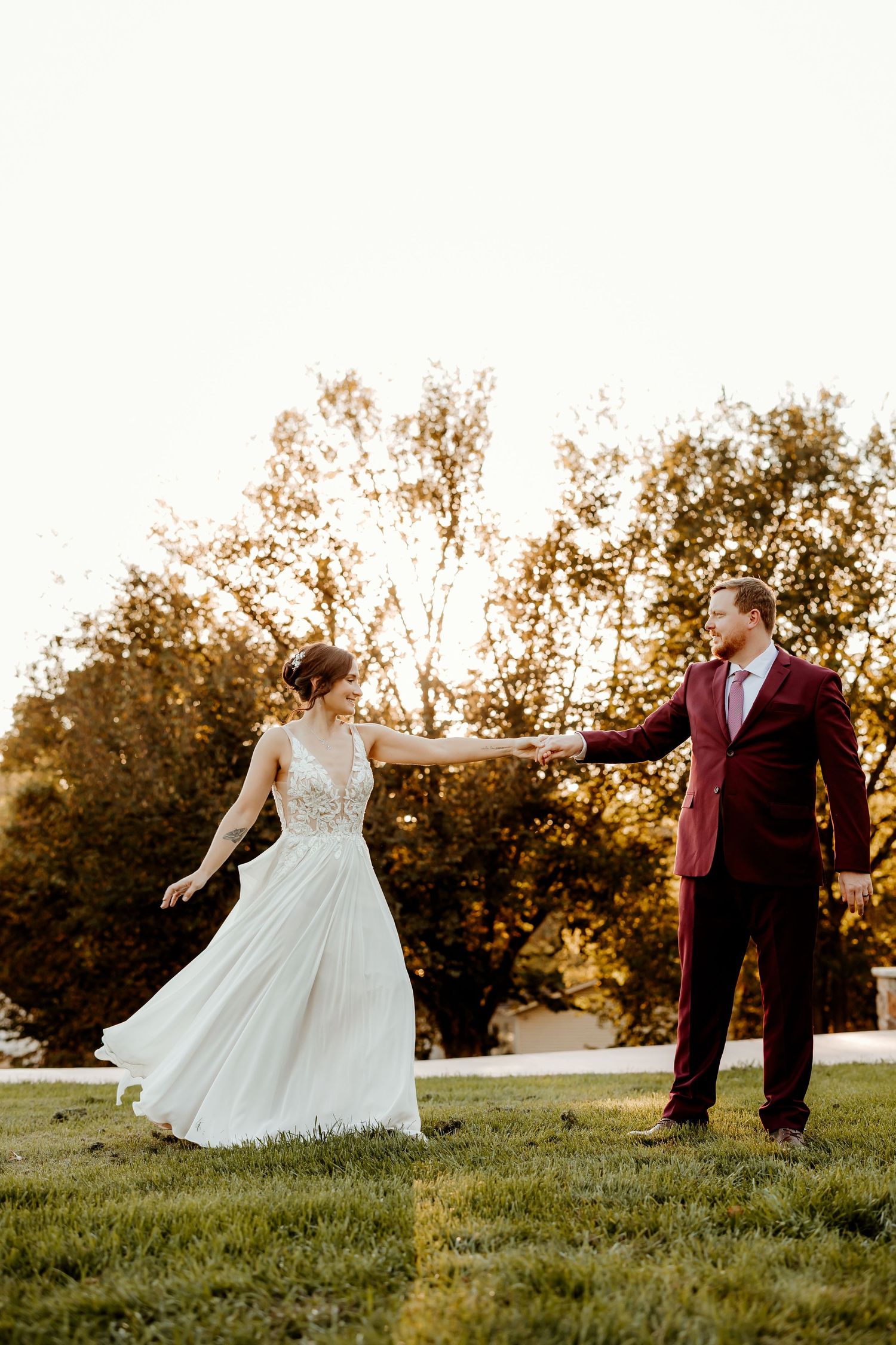 Bride in a flowing white lace gown holds hands with groom in a burgundy suit while dancing on a grassy lawn at golden hour.