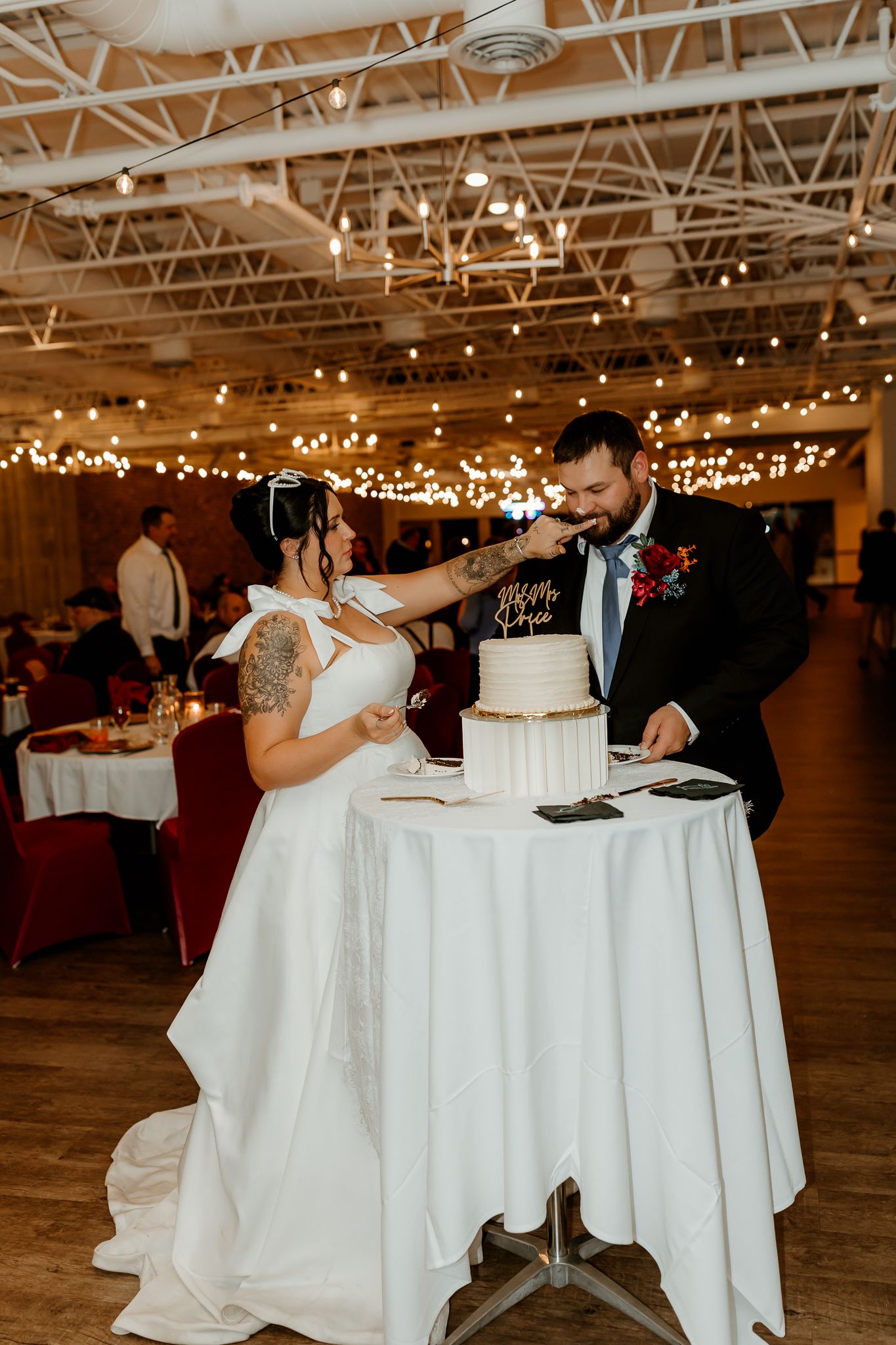 Bride in a white gown and groom in a dark suit cut their white tiered wedding cake at a reception with string lights overhead.