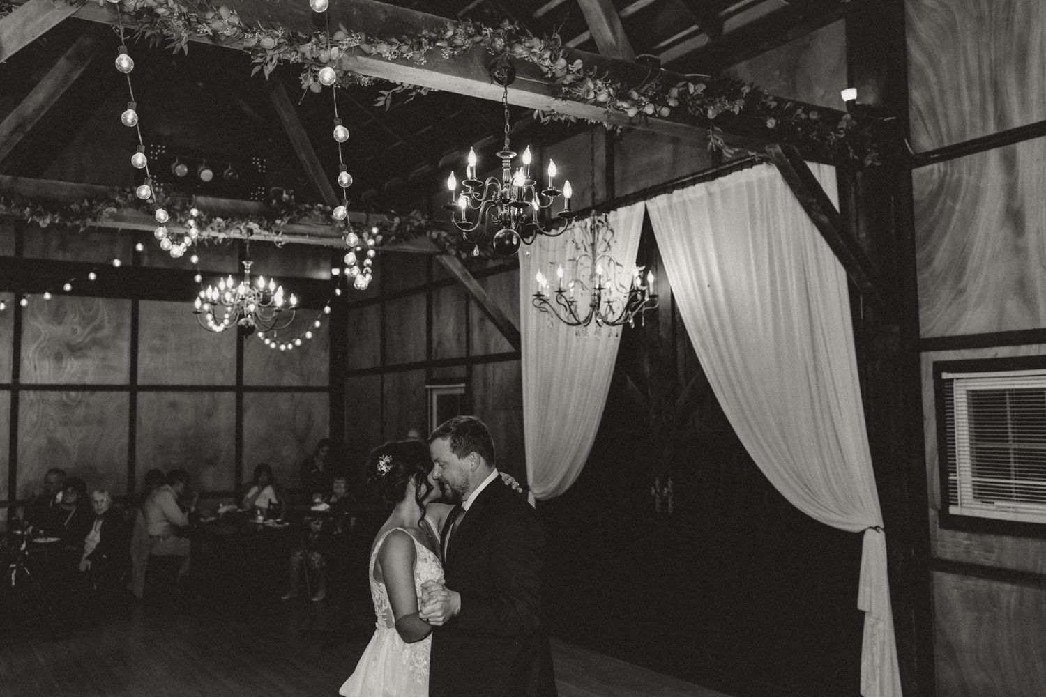 Bride and groom share a first dance in a rustic barn wedding venue with chandeliers, string lights, and draped white curtains.