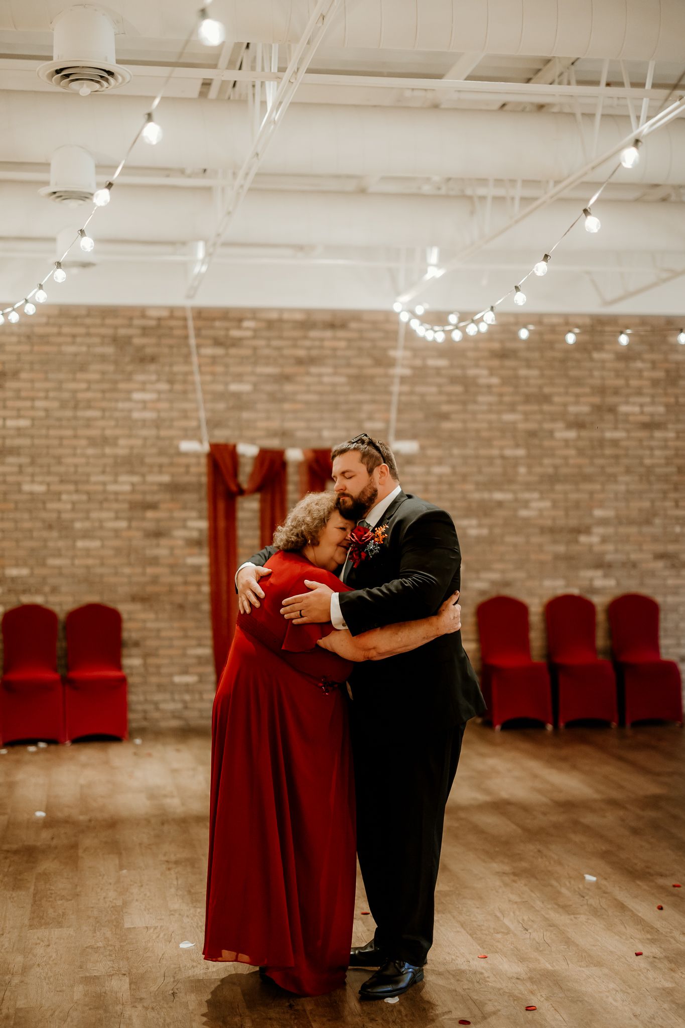 A man in a black suit embraces a woman in a long red dress on a wood floor in a brick venue with string lights and red chairs.